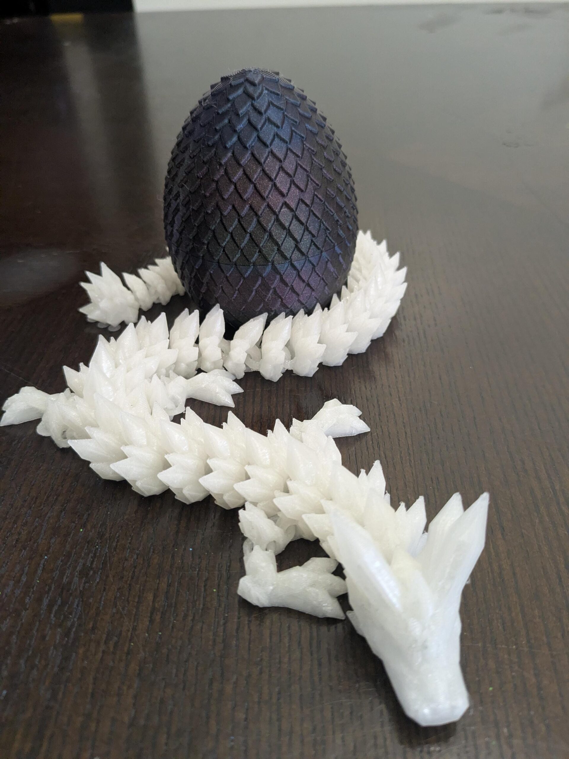 May include: A fantasy-themed sculpture featuring a dark, textured egg with iridescent highlights, resting on a white, spiky, crystal-like base. A white dragon with a crystal-like body extends from the base, creating a unique decorative piece.