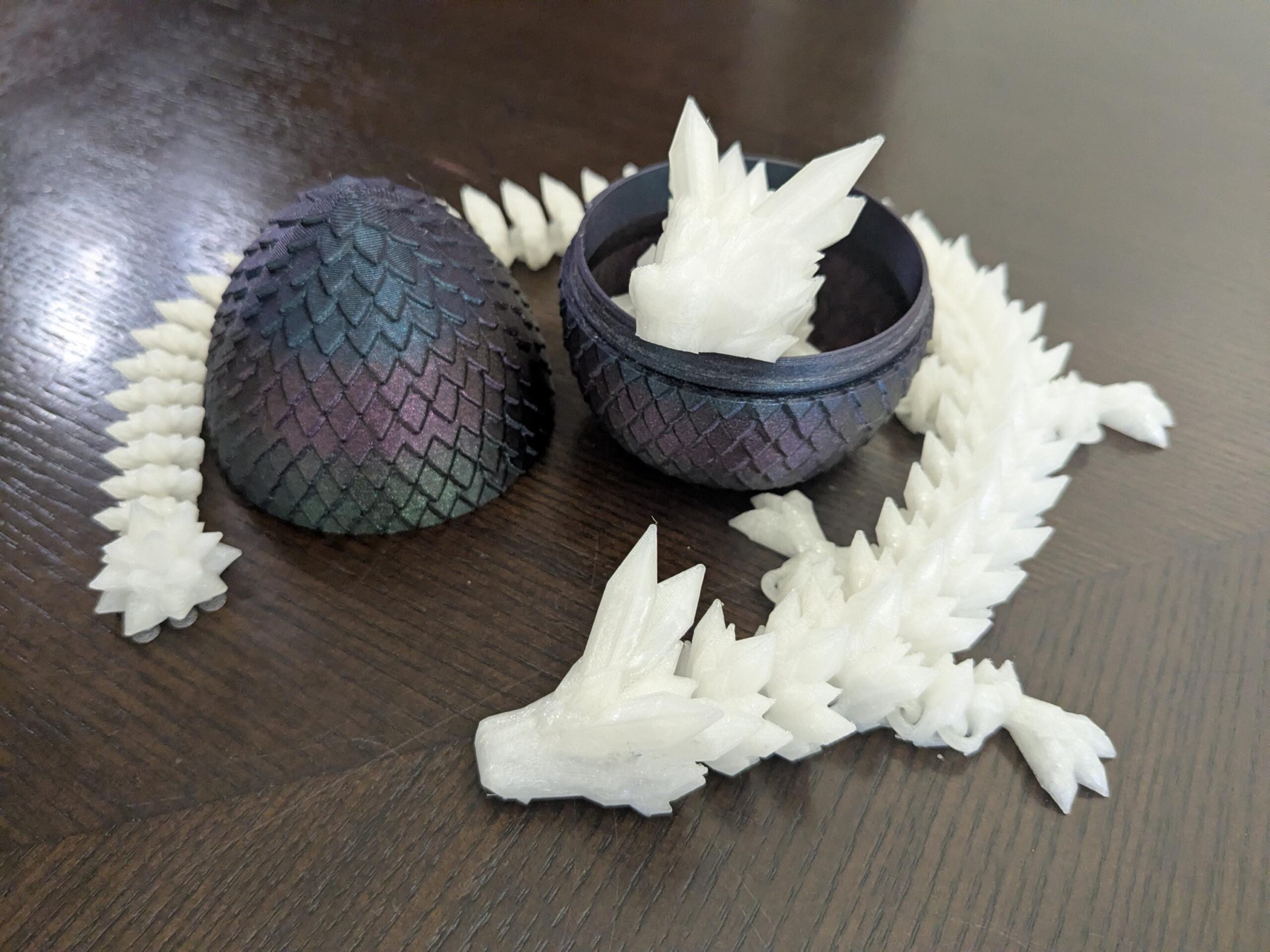 May include: A 3D-printed dragon toy with a segmented white body and head, emerging from a dark blue and purple egg. The egg is open, revealing the dragon inside. The eggshell has a textured, scale-like pattern.