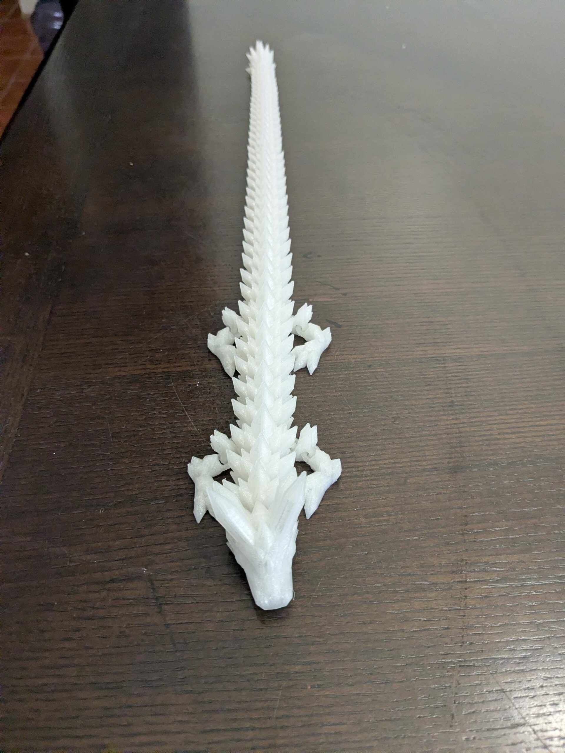 May include: A white, 3D-printed dragon figurine. The dragon is depicted in a top-down view, with a long, spiky tail and detailed scales. The figurine is made of a solid white material and rests on a dark brown wooden surface.