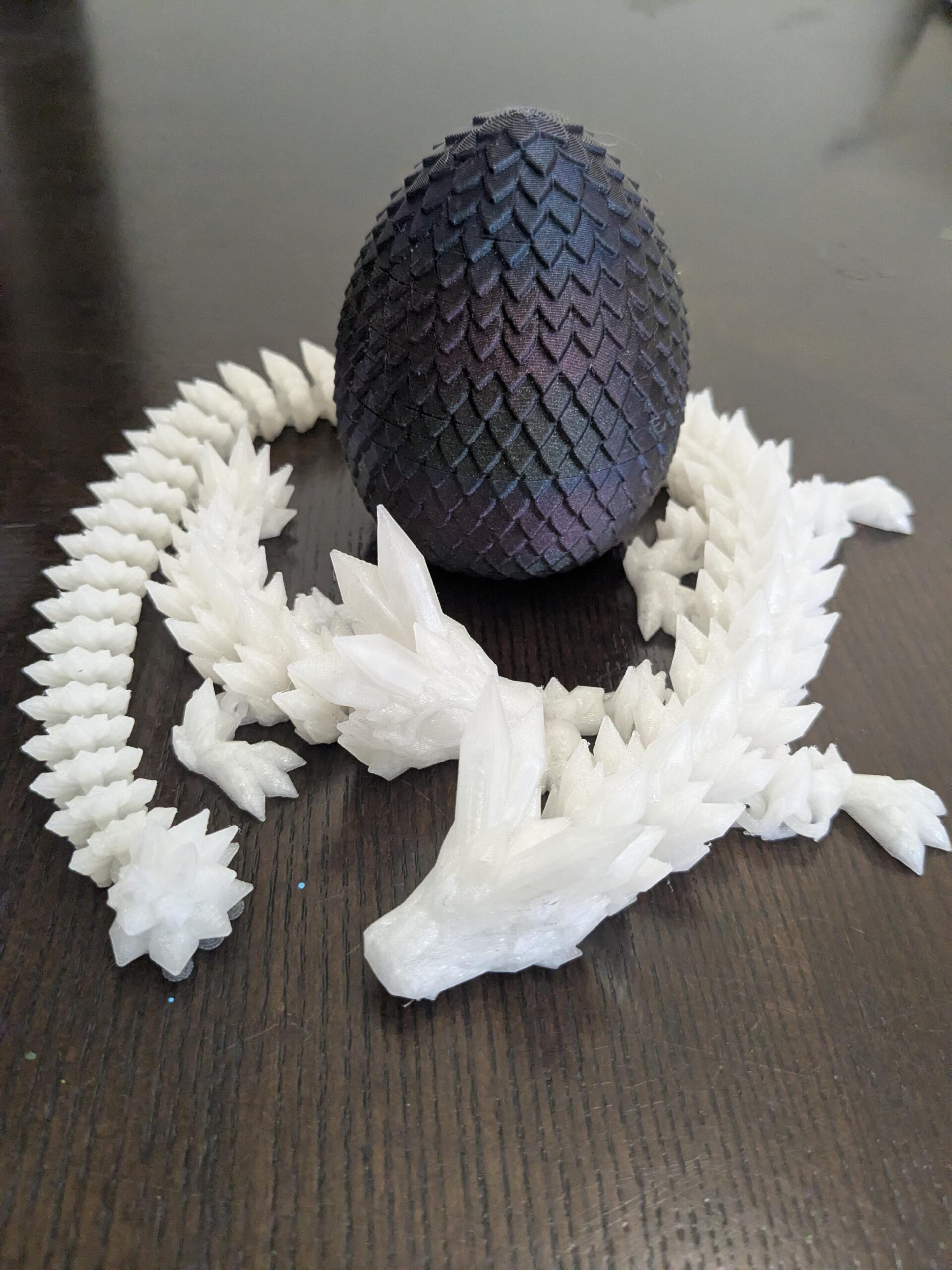 May include: A 3D-printed dragon egg with a textured, iridescent finish in shades of blue, purple, and black. A white, articulated dragon figure surrounds the egg, with a spiky, crystalline design.