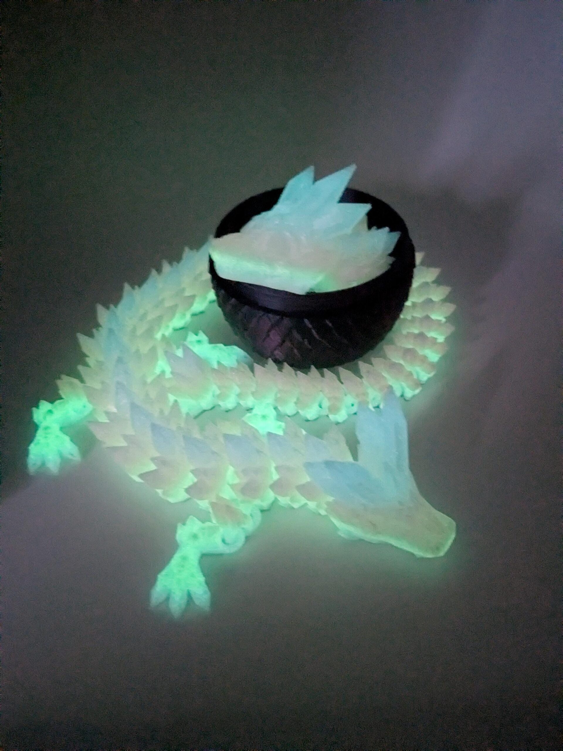 May include: A glowing, light blue and white dragon sculpture with a black bowl-shaped object on its back. The dragon's scales and the object's contents emit a soft, green light. The sculpture is on a gray background.