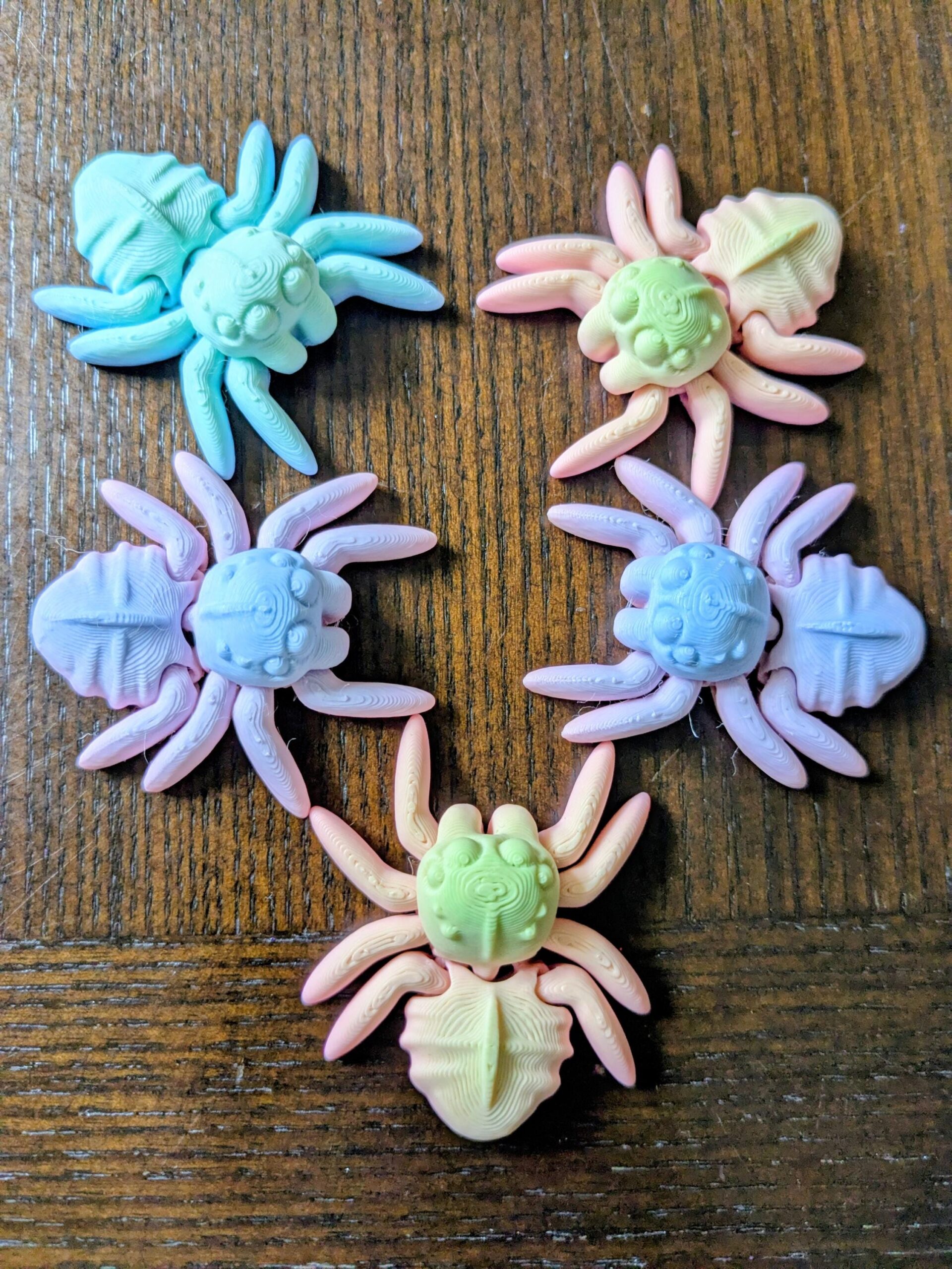 May include: Six colorful spider-shaped ornaments in pastel shades of blue, pink, yellow, and green. The spiders have detailed bodies and legs, and are arranged on a wooden surface. These decorative ornaments are ideal for Halloween or other themed crafts.