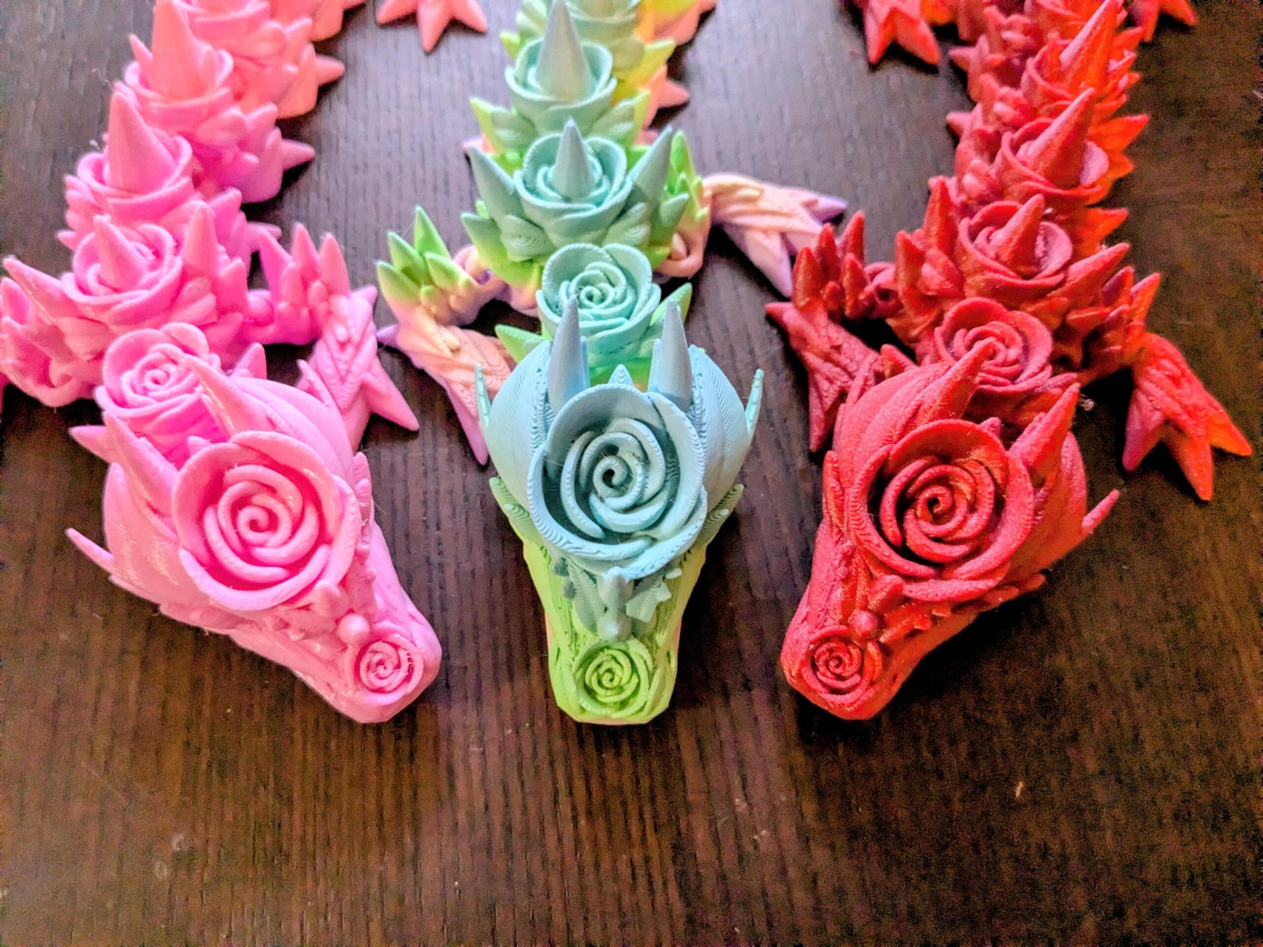 May include: Three colorful dragon sculptures with rose-like details. One is pink, one is red, and one is a gradient of blue and green. The dragons are on a dark wooden surface.