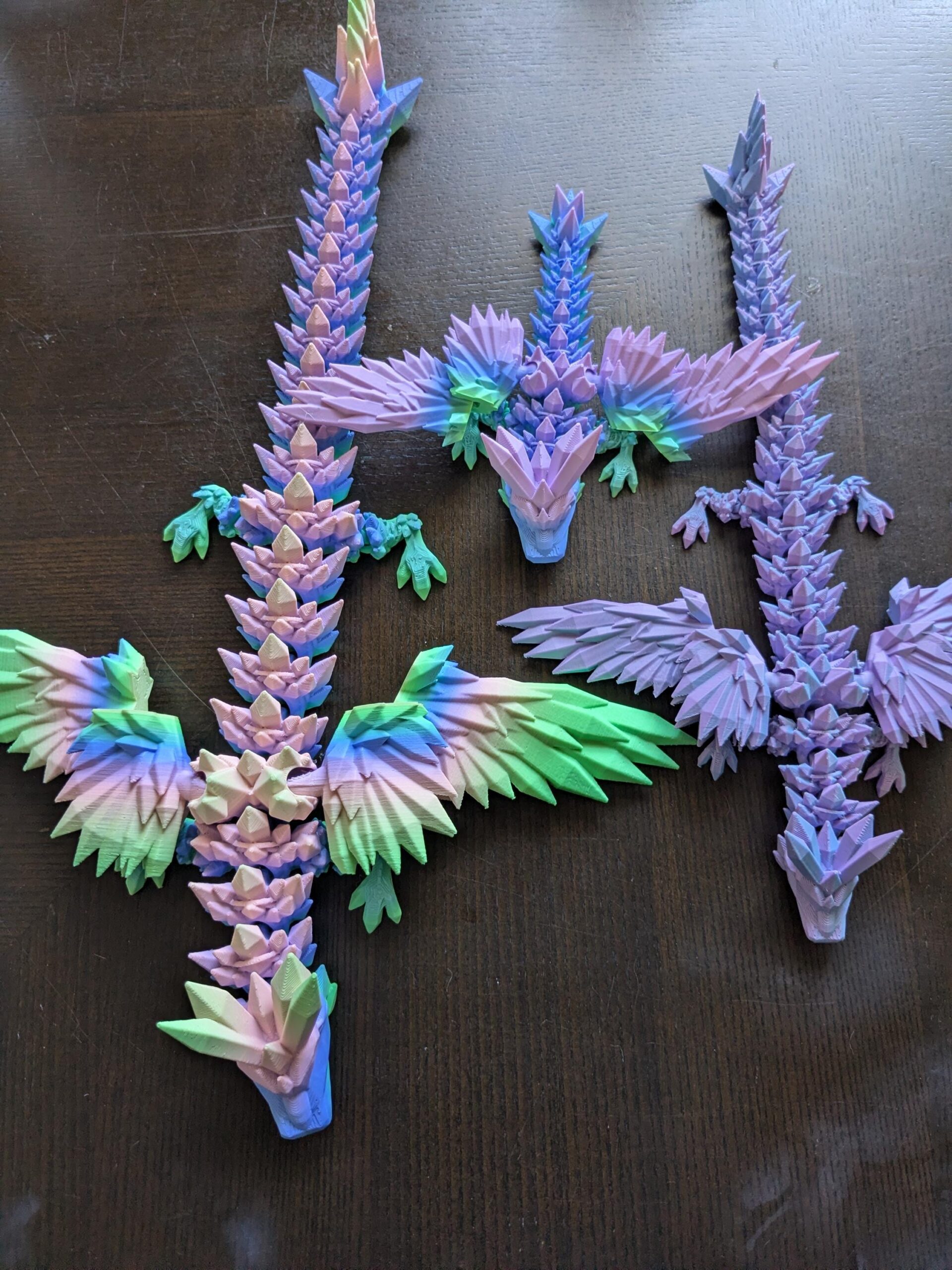 May include: Three 3D-printed dragon figurines in shades of purple, blue, and green. The dragons have detailed, spiky bodies, outstretched wings, and are posed on a dark brown surface. The wings and bodies have a gradient color effect.