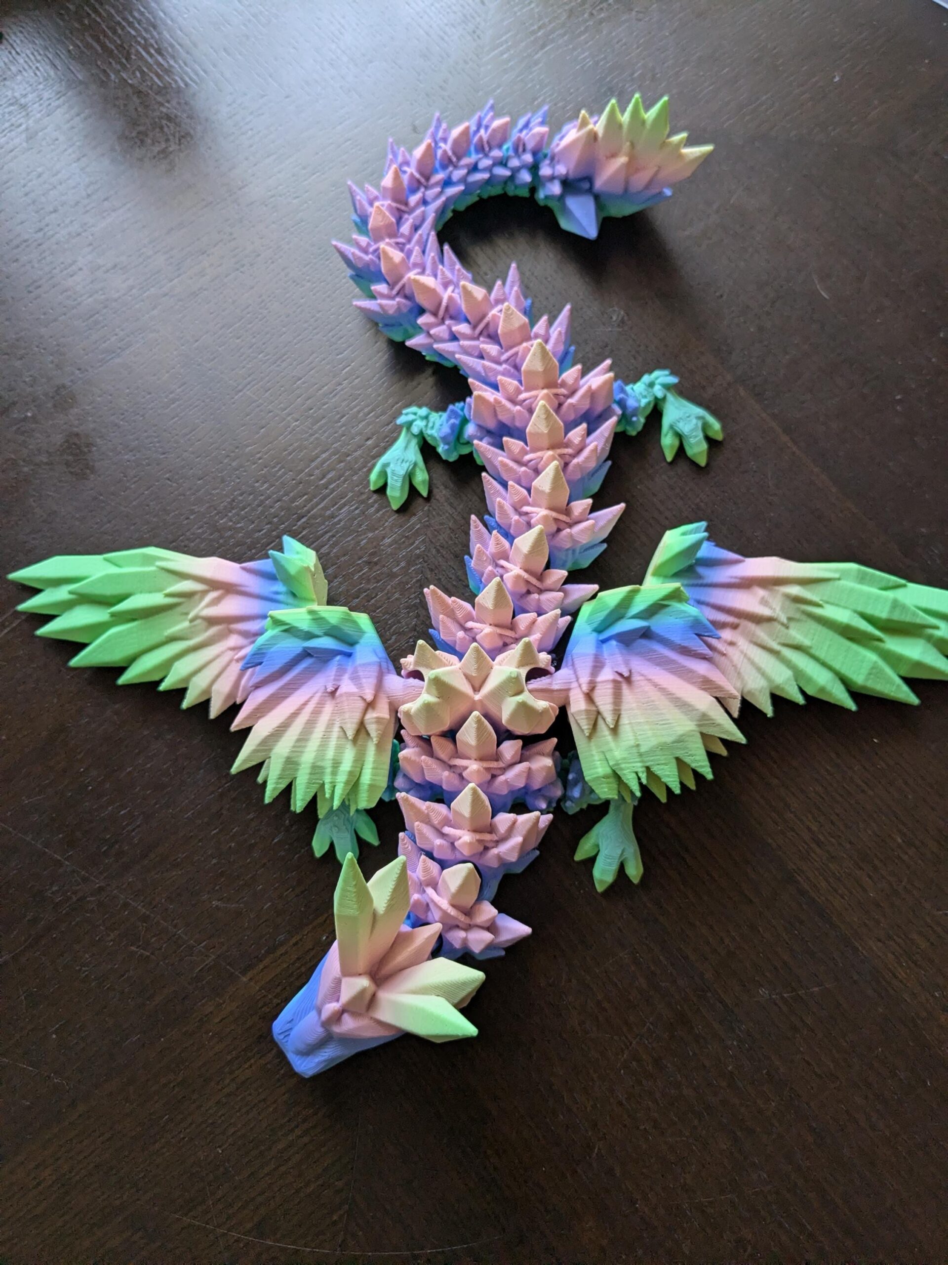 May include: A colorful, 3D-printed dragon figurine with a geometric design. The dragon features a segmented body, wings, and a spiky spine, with a color palette of blue, green, pink, and purple. The wings are spread, and the dragon is posed on a dark surface.