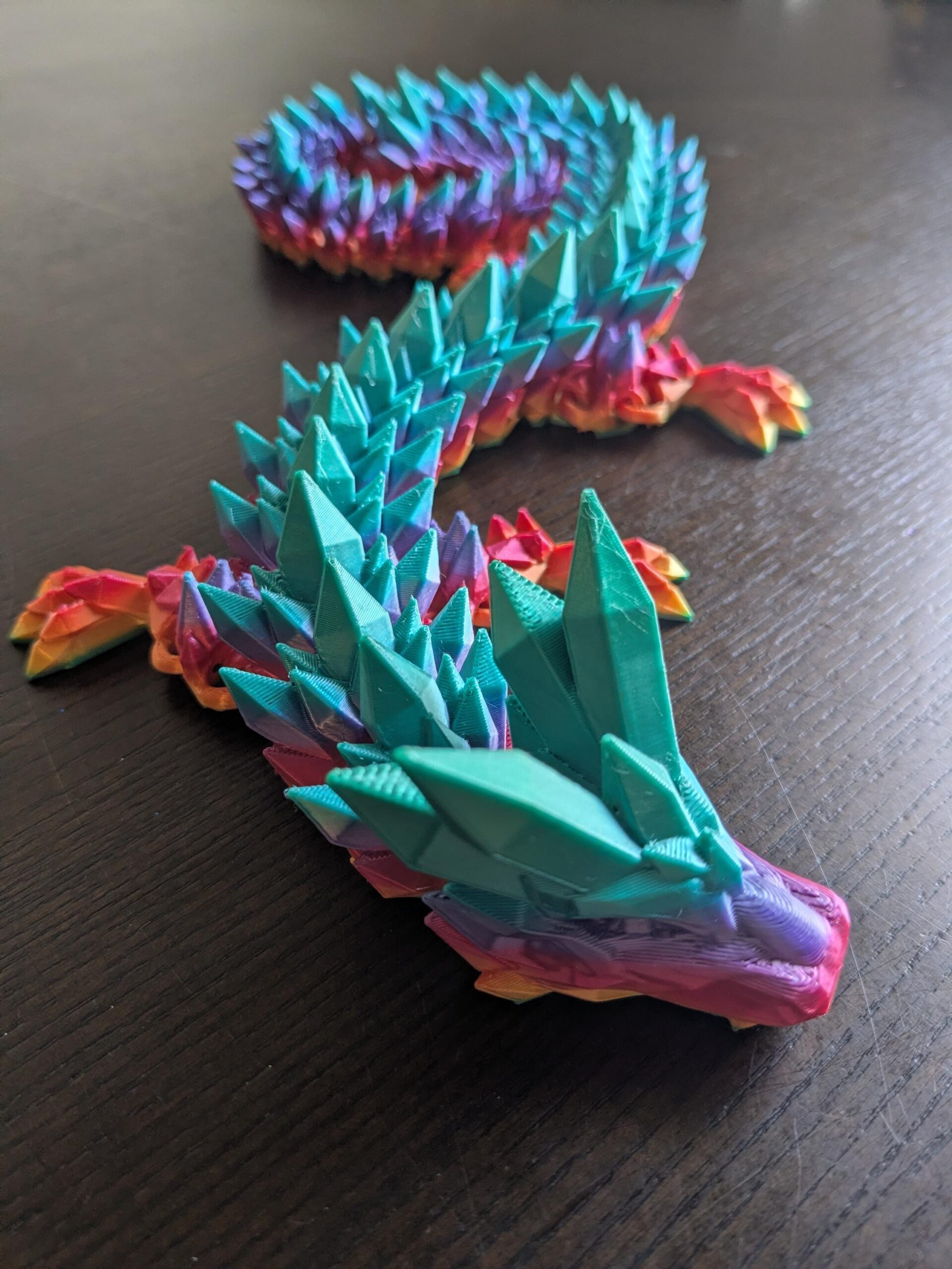 May include: A colorful, articulated dragon figurine with a crystal-like design. The dragon's body transitions through shades of red, orange, yellow, purple, and blue. The head and spine feature prominent, teal-colored crystal spikes. The dragon is posed on a dark surface.