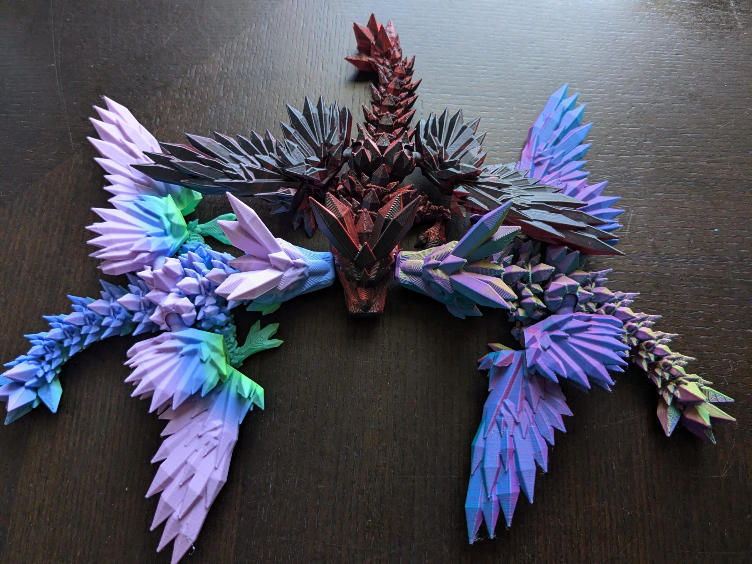 May include: A multi-colored, geometric dragon sculpture with a central body in shades of red and black, and wings in blue, purple, and green. The dragon is made of angular, crystalline shapes, creating a unique and modern aesthetic. The sculpture is displayed on a dark surface.