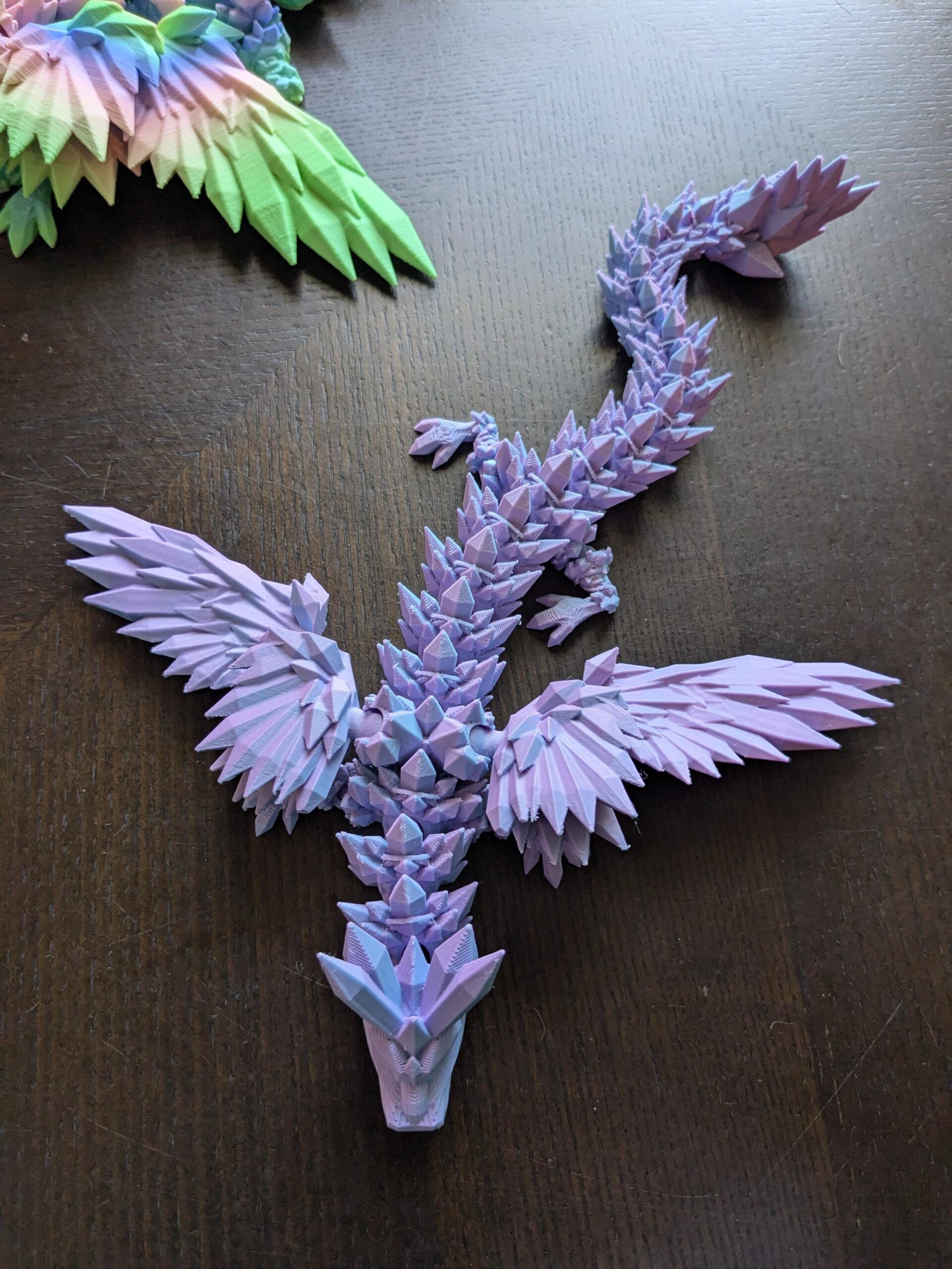 May include: A 3D-printed dragon figurine in shades of purple, blue, and pink. The dragon has detailed wings and a spiky spine. The figurine is a fantasy-themed decorative item.