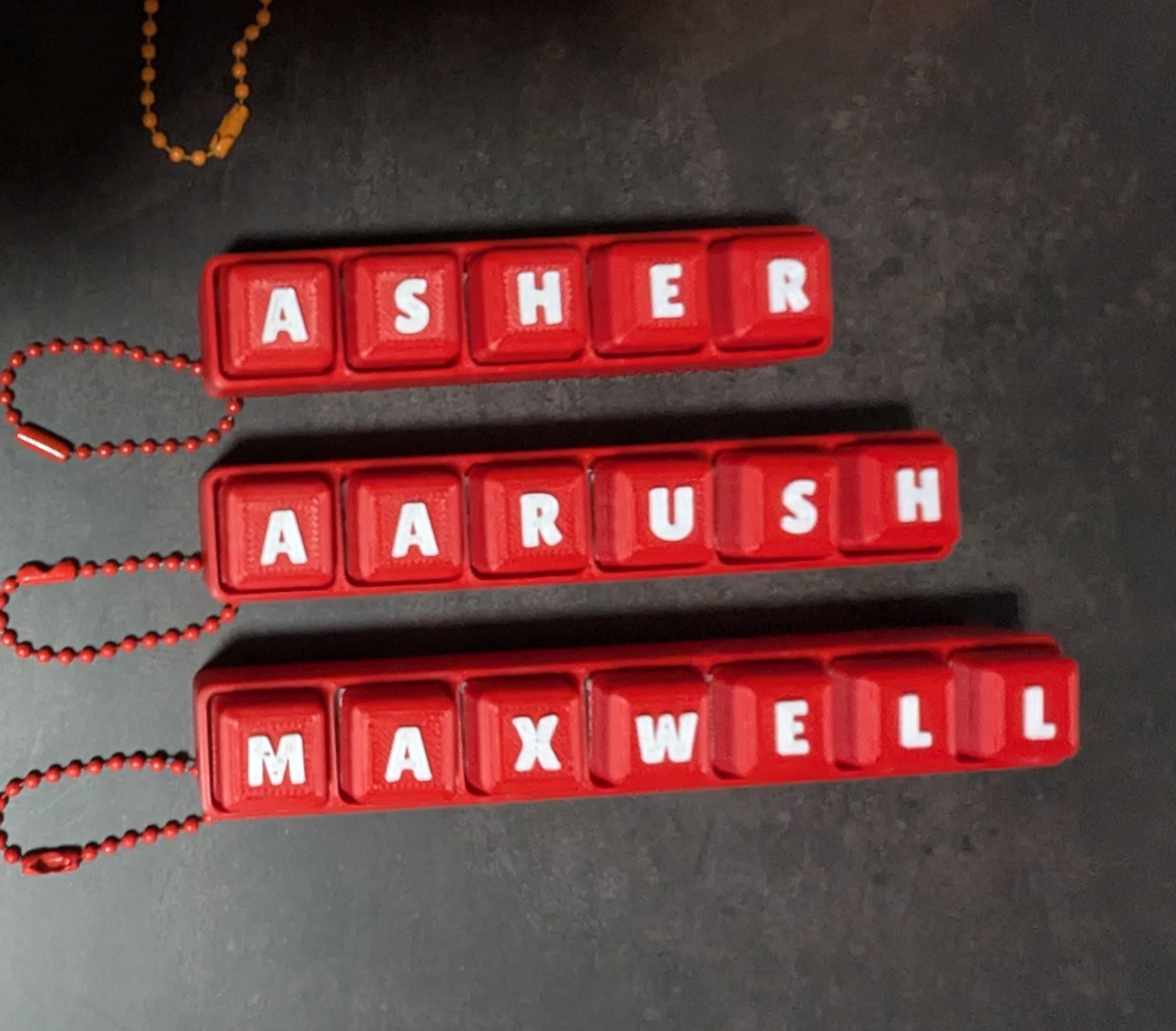 May include: Three red keychains with white letters spelling out the names "Asher", "Aarush", and "Maxwell". Each keychain has a red ball chain attached. The keychains are displayed on a dark gray surface.