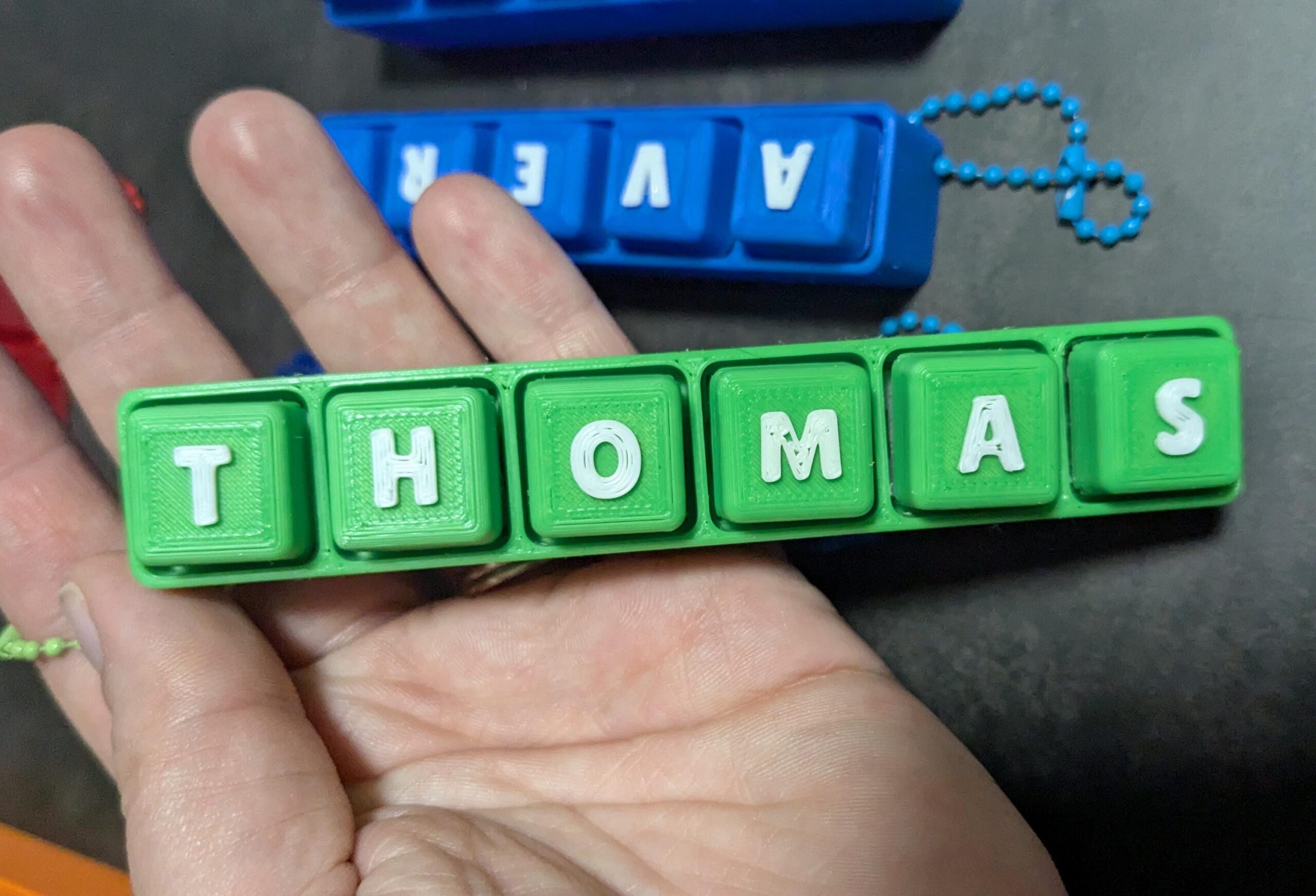 May include: A green keychain spelling "THOMAS" in white letters. The keychain is made of individual square blocks. A blue keychain spelling "AVER" is visible in the background.