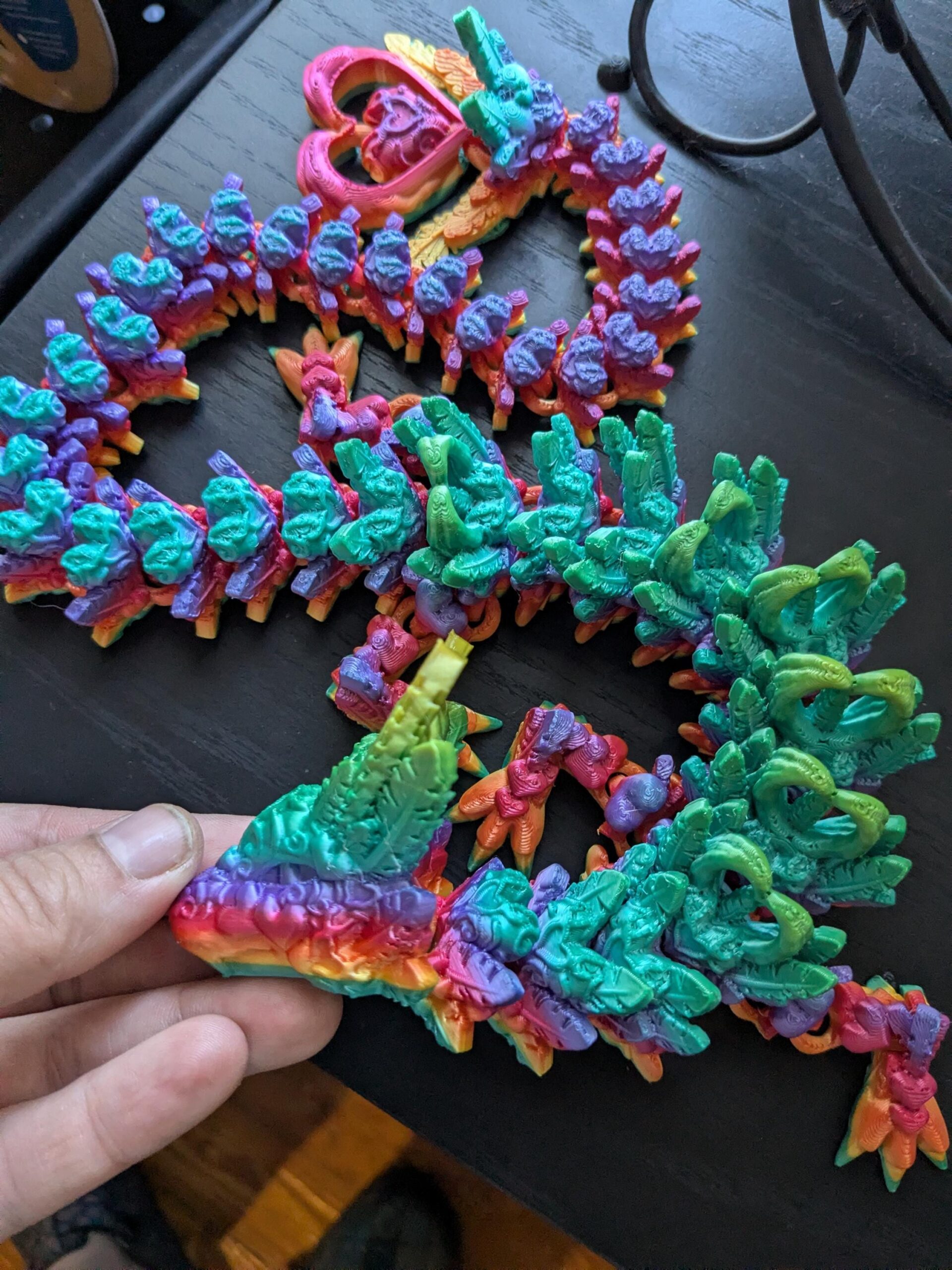 May include: A colorful, articulated dragon toy in shades of rainbow, purple, green, and blue. The dragon is 3D-printed and has a segmented body, with a heart-shaped design at the top. The toy is held by a person.