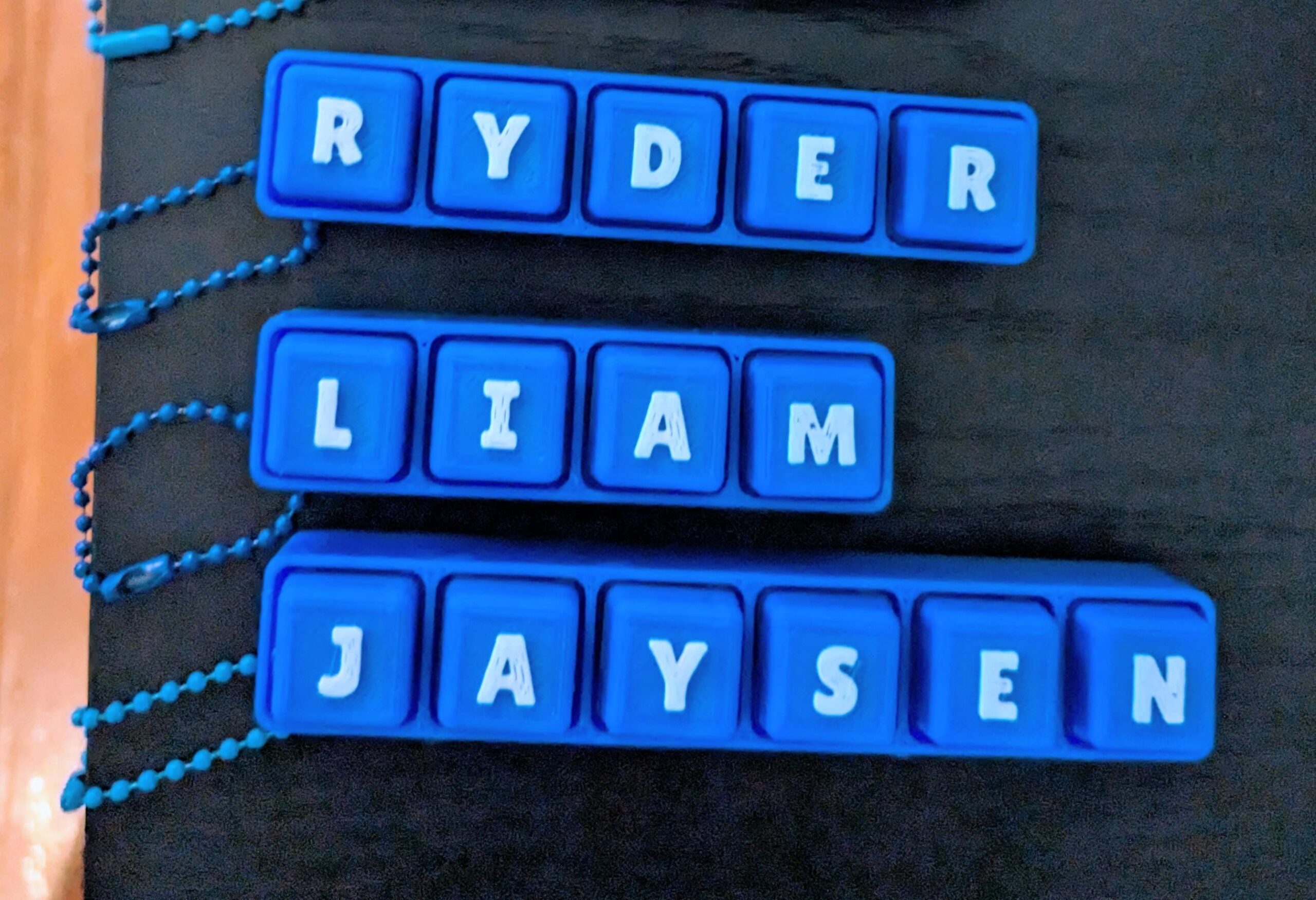 May include: Three blue name tags spelling out "RYDER", "LIAM", and "JAYSEN" in white capital letters. Each name is on a separate blue rectangular tag with a chain attached.