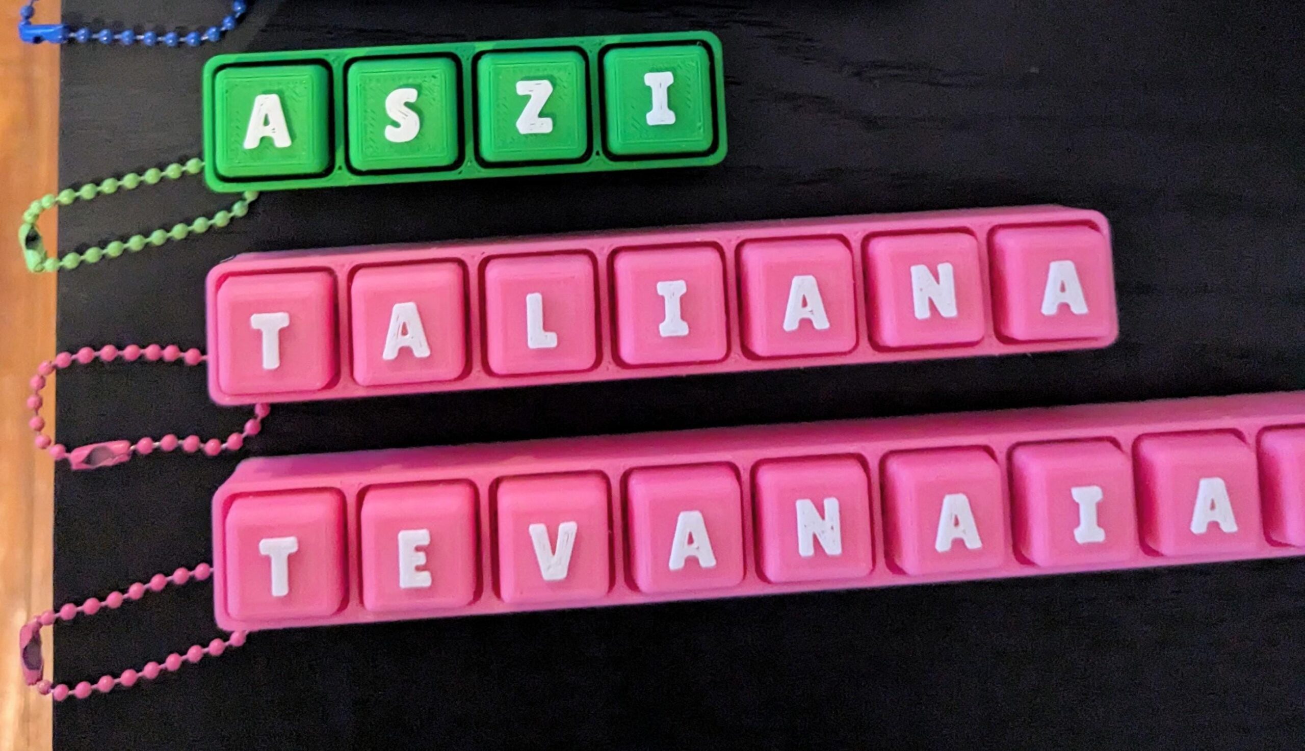 May include: Three personalized name keychains. One green keychain spells out "ASZI" in white letters. Two pink keychains spell out "TALIANA" and "TEVANAIA" in white letters. Each keychain has a matching colored chain.