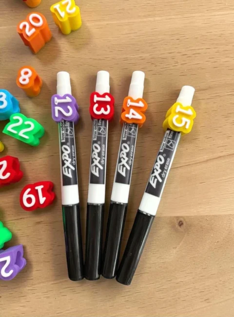 May include: Four black Expo dry erase markers with white caps, each featuring a colorful silicone number topper. The markers are labeled 12 in purple, 13 in red, 14 in orange, and 15 in yellow. Scattered around are additional silicone number pieces.