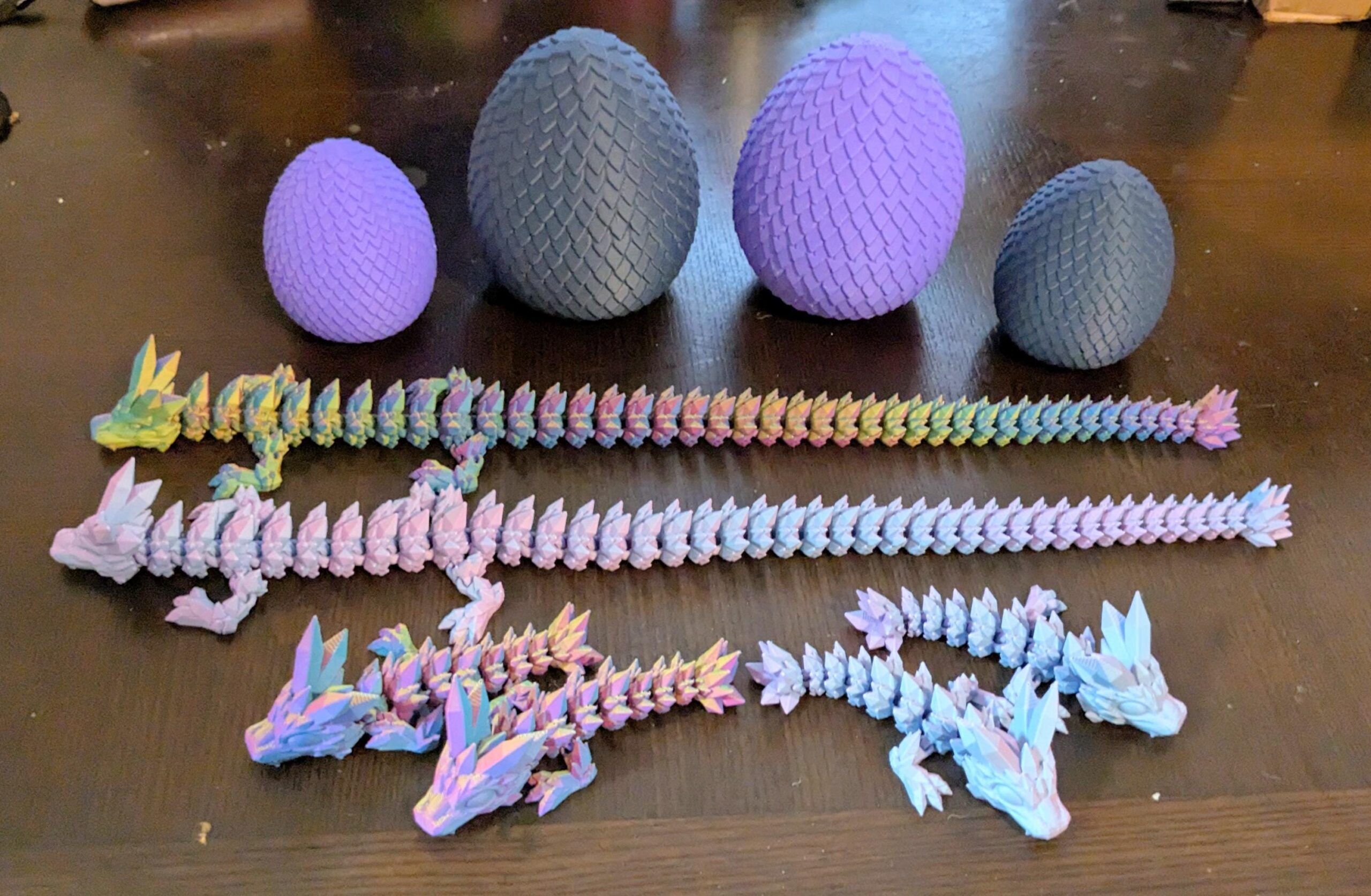 May include: A collection of 3D-printed dragon toys and textured eggs in various sizes and colors. The dragons are articulated and multi-colored, while the eggs are purple and gray.
