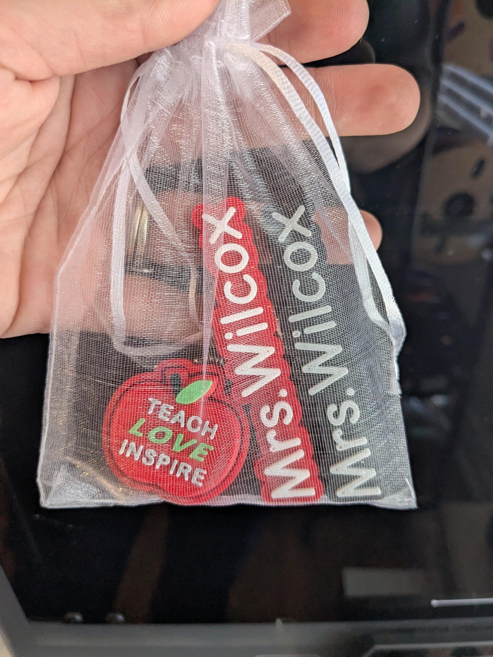 May include: A clear, sheer organza gift bag containing three items. One is a red apple-shaped charm with the words "Teach Love Inspire". Two rectangular charms read "Mrs. Wilcox" in white on red and black backgrounds.