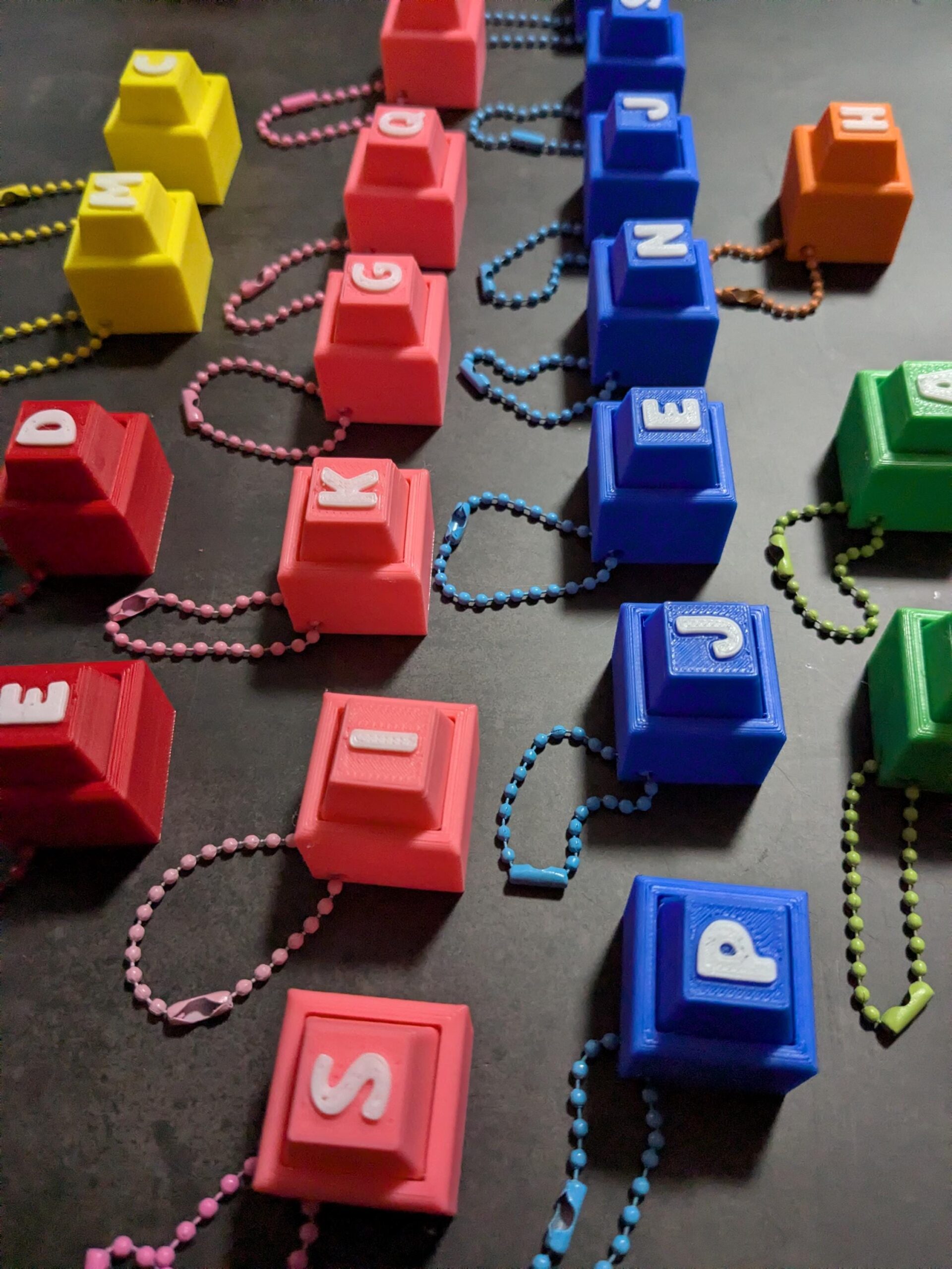 May include: A collection of colorful, 3D-printed keychains. Each keychain is a cube with a raised square top, featuring a different letter. The keychains are in various colors, including red, yellow, blue, green, and orange, and are attached to beaded chains.