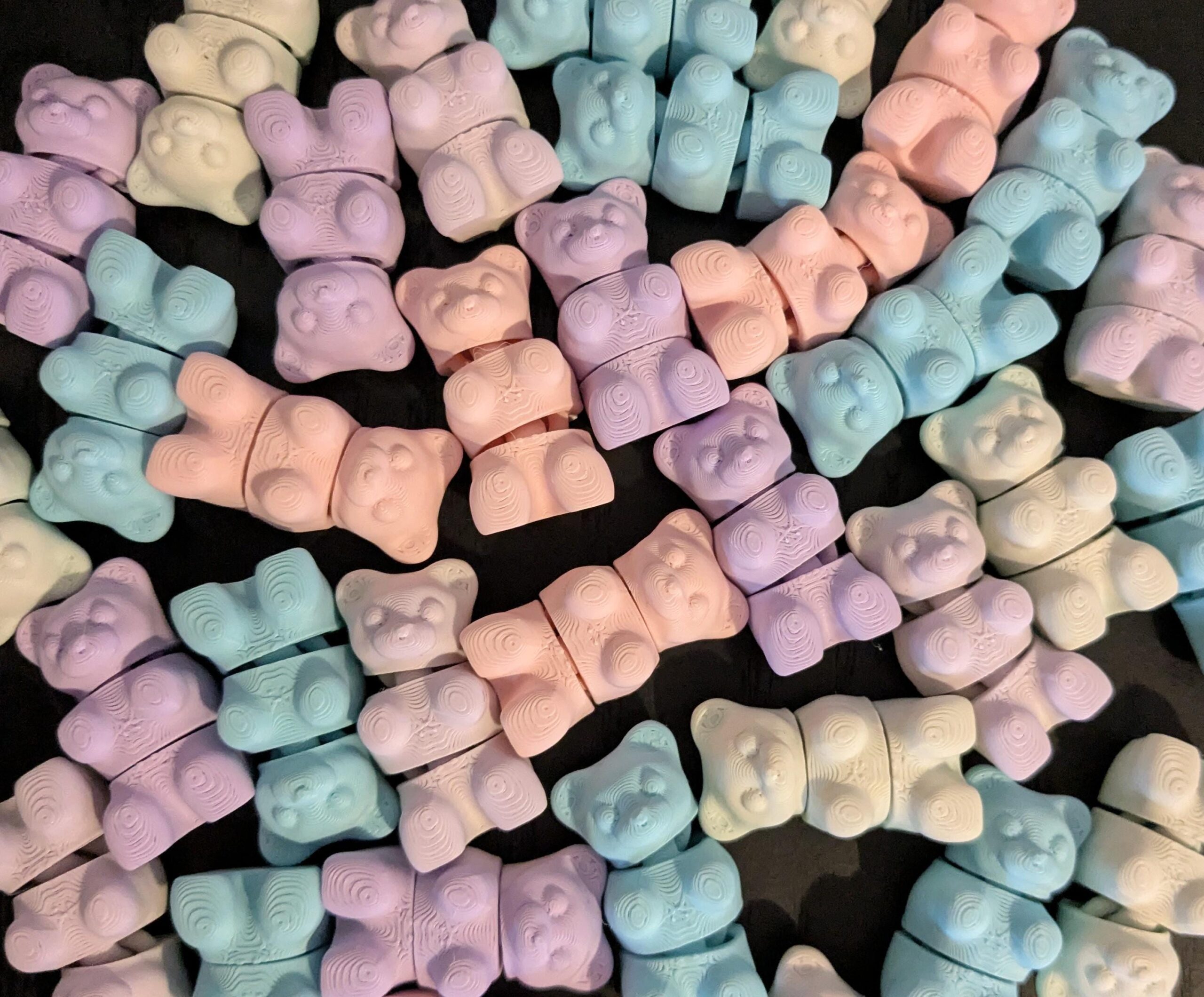 May include: A collection of pastel-colored gummy bear-shaped erasers in shades of pink, purple, blue, and cream. The erasers are segmented into two pieces, with a detailed bear design. The background is a dark surface.