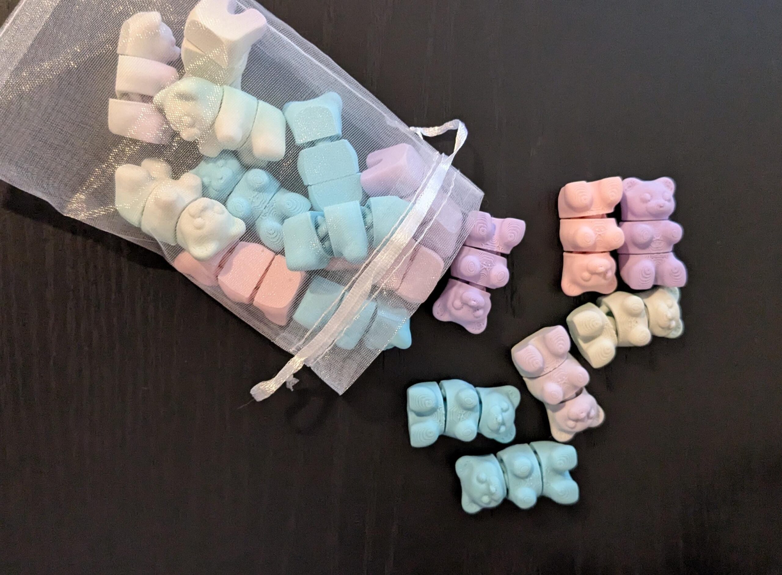 May include: A collection of pastel-colored gummy bear candies in a small, white mesh bag. The bears are pink, blue, purple, and white. Some are stacked in small piles on a dark surface.