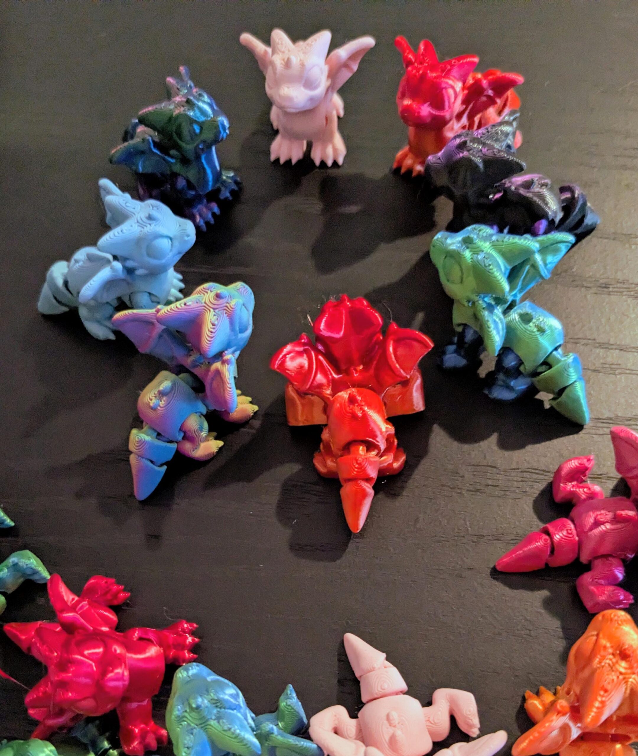 May include: A collection of colorful, 3D-printed dragon figurines in various colors, including red, blue, green, and pink. The dragons have detailed wings, horns, and claws, and are arranged in a circular pattern on a dark surface.