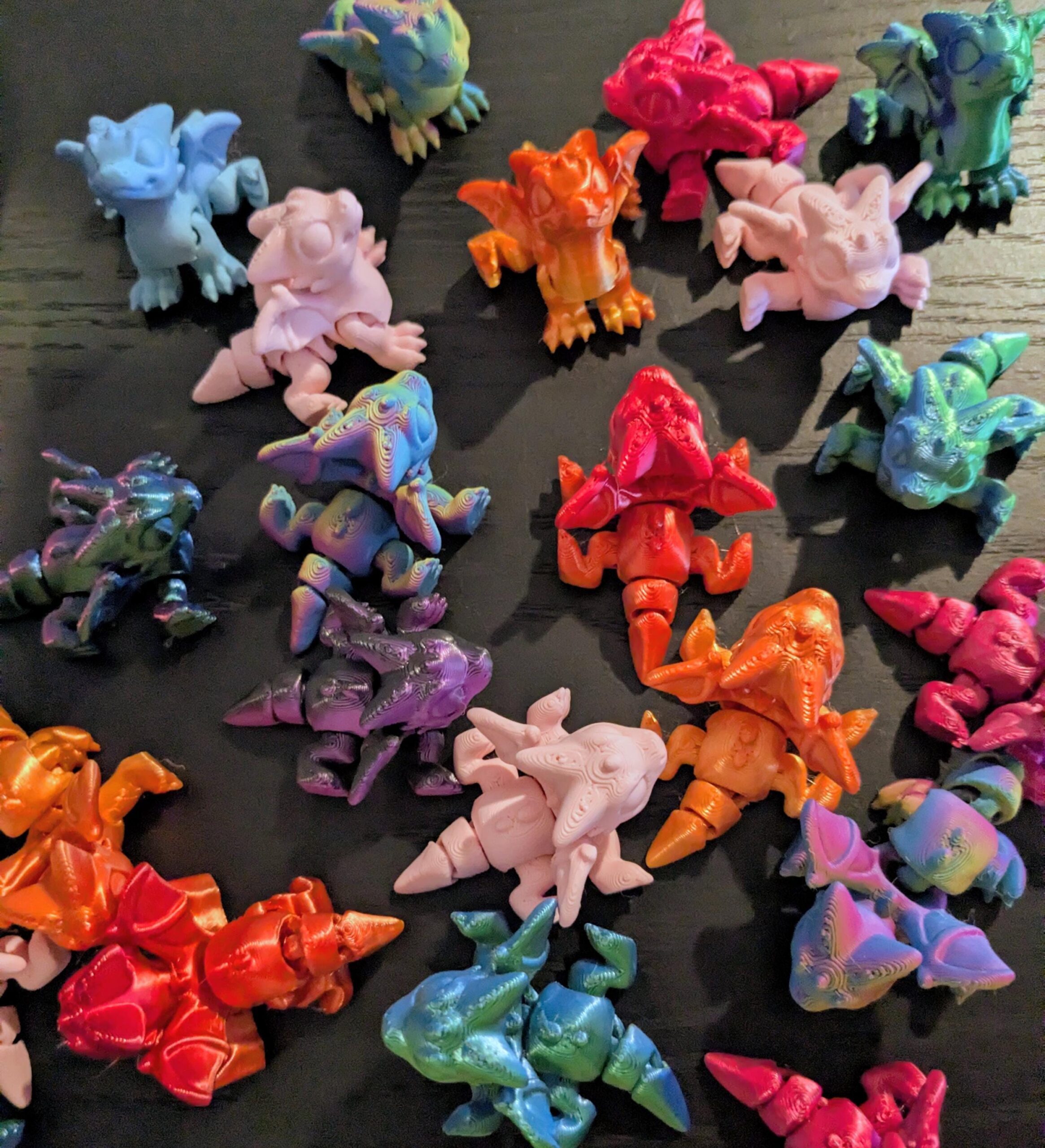 May include: A collection of colorful, articulated dragon figurines in various shades including blue, pink, orange, and green. The dragons have detailed wings, legs, and tails, and are arranged on a dark surface. The figurines appear to be 3D-printed.