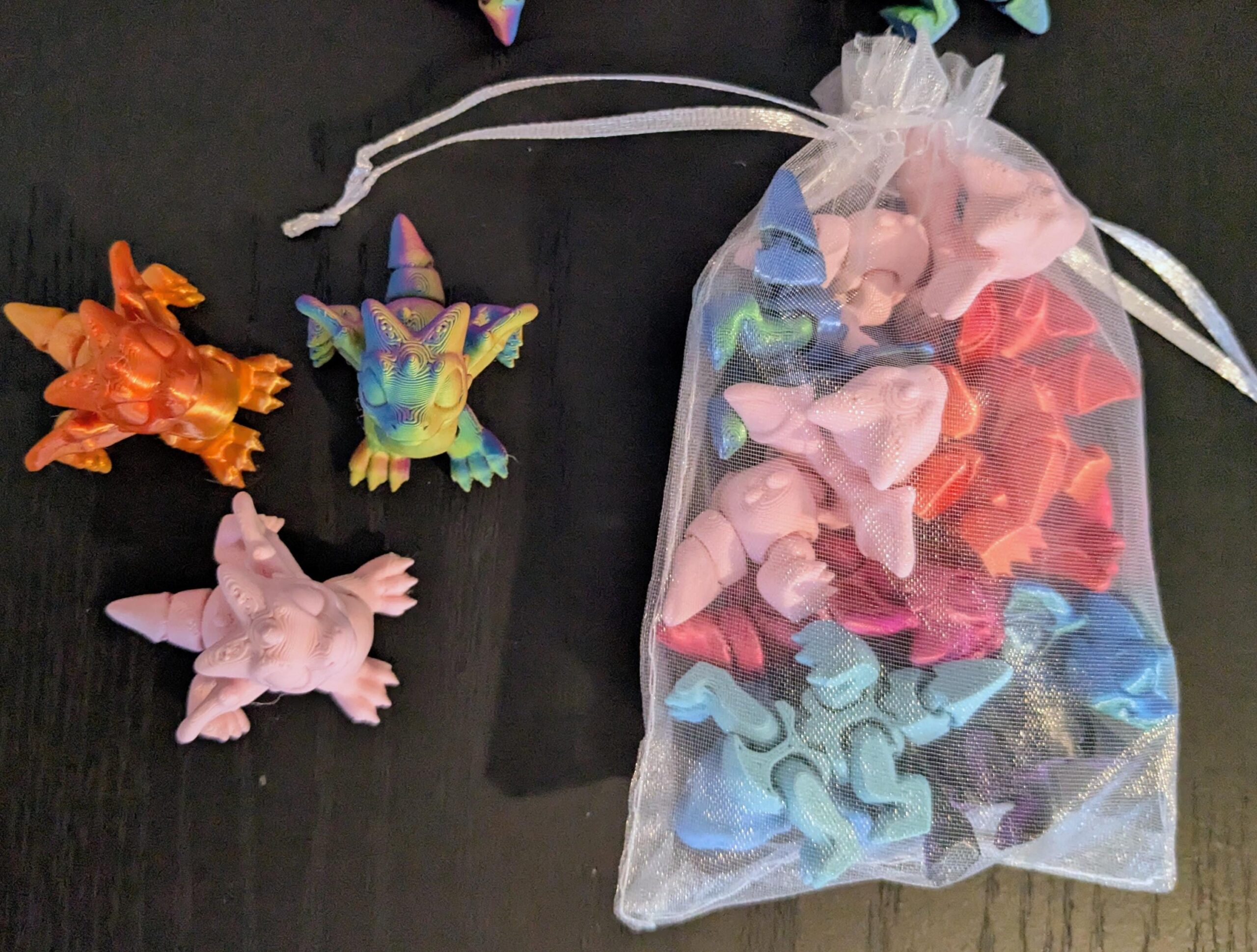 May include: A collection of colorful, miniature dinosaur figurines. Three figurines are displayed outside a sheer white drawstring bag filled with more figurines. The dinosaurs are various colors, including orange, pink, blue, and rainbow.