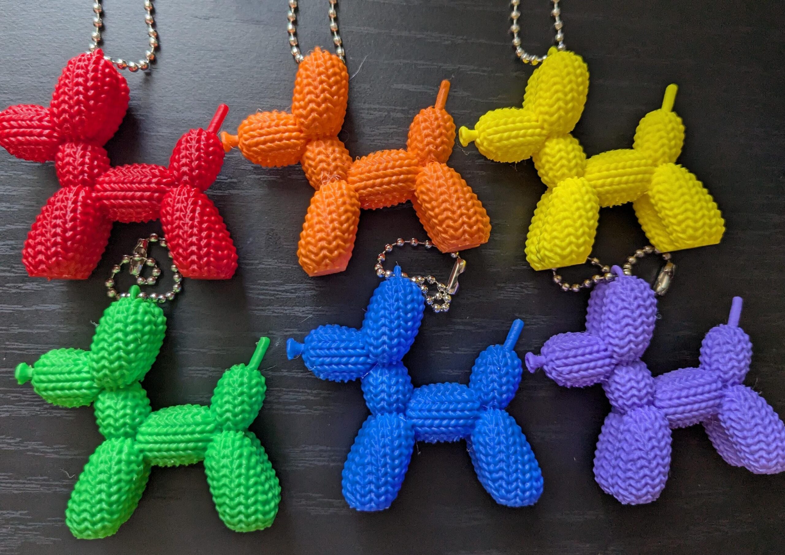 May include: A collection of six colorful balloon dog keychains in red, orange, yellow, green, blue, and purple. Each keychain is attached to a silver ball chain. The dog figures have a textured, knitted appearance.