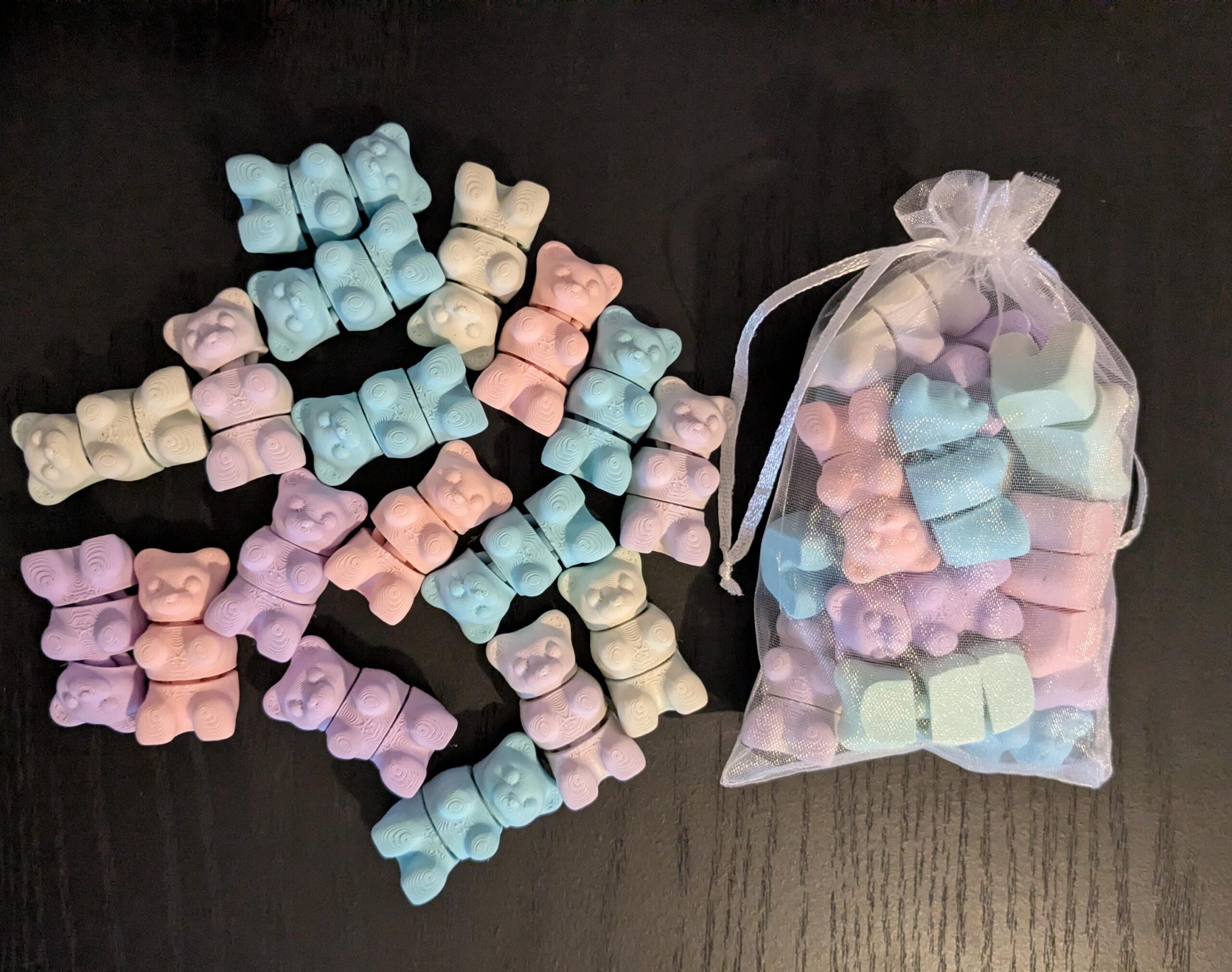 May include: A collection of pastel-colored gummy bear-shaped soaps in shades of pink, blue, purple, and cream. Some are scattered on a dark surface, while others are contained in a sheer, drawstring bag. Each soap is approximately 1 inch in size.