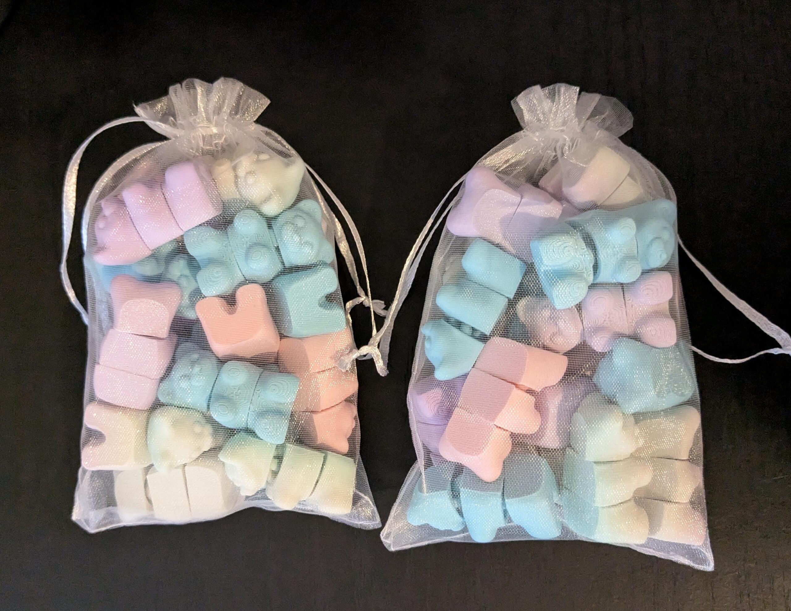 May include: Two small, sheer white drawstring bags filled with pastel-colored gummy bear-shaped candies. The candies are in shades of pink, blue, purple, and white. The bags are tied with white satin ribbon.
