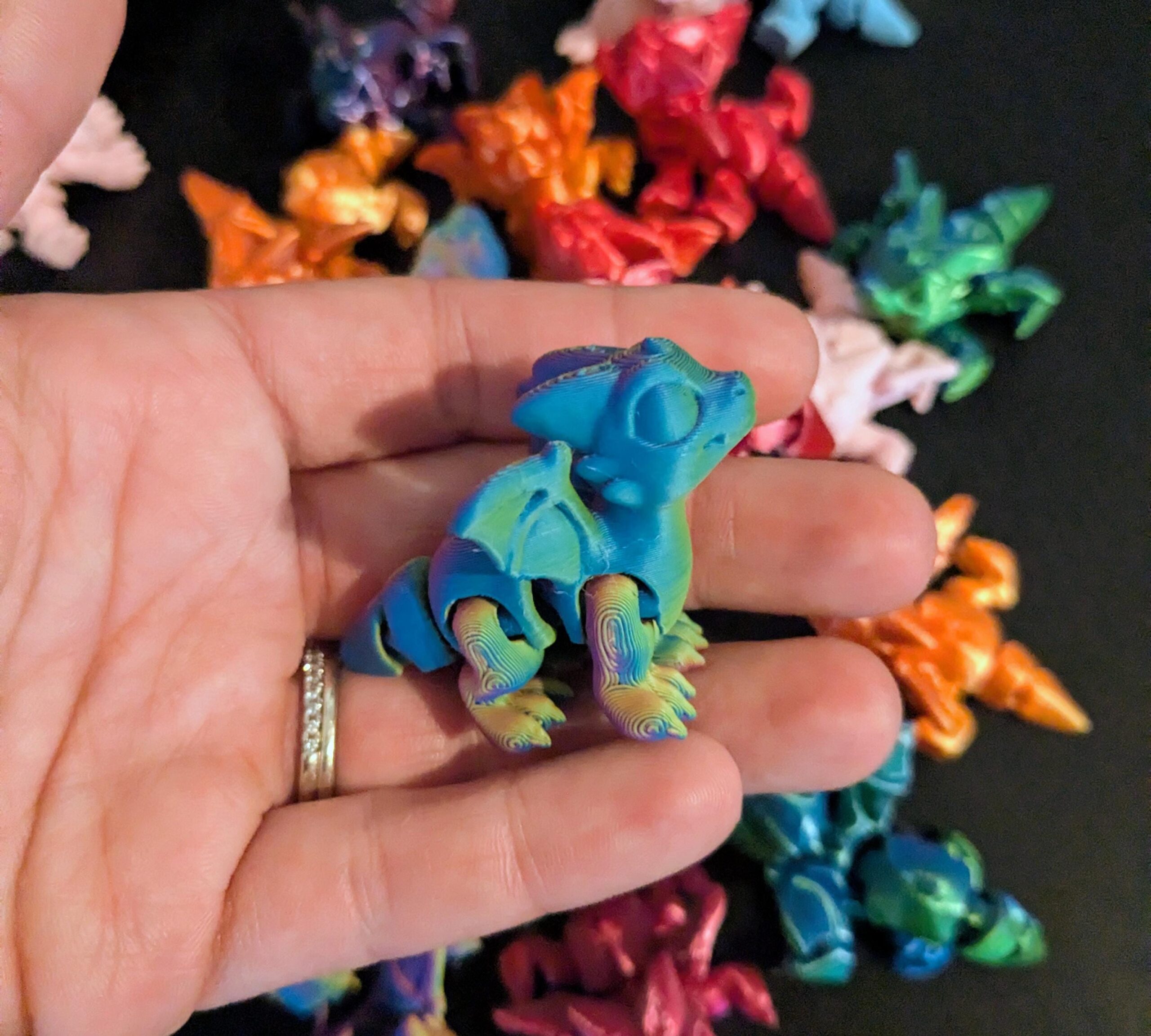 May include: A blue and green articulated dragon figurine with iridescent coloring. The dragon is held in a hand, with other colorful dragon figurines in the background. The dragon has wings, legs, and a tail.