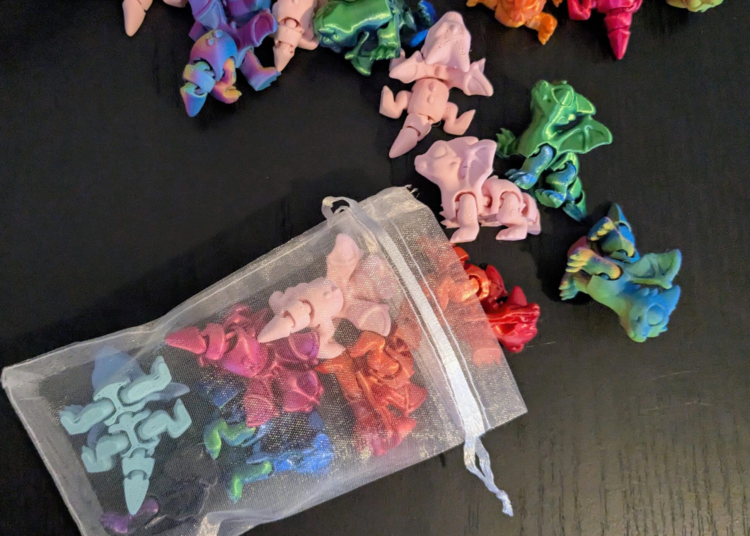May include: A collection of colorful, articulated dragon figurines in various shades of pink, blue, green, and orange. Some dragons are loose, while others are in a sheer drawstring bag. The background is a dark surface.