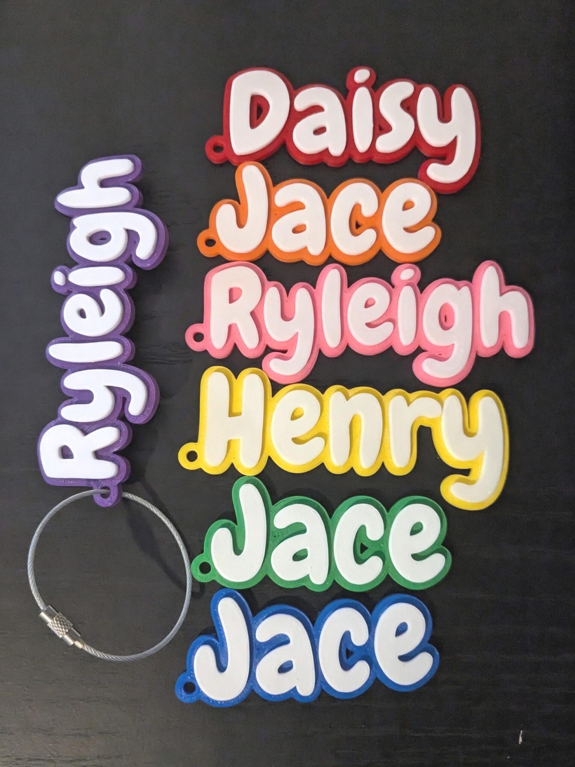 May include: A collection of six custom name tags featuring white raised lettering on colorful backgrounds. The names include Daisy in red, Jace in orange, Ryleigh in pink, Henry in yellow, Jace in green, and Jace in blue. One purple Ryleigh tag includes a silver metal cable loop.