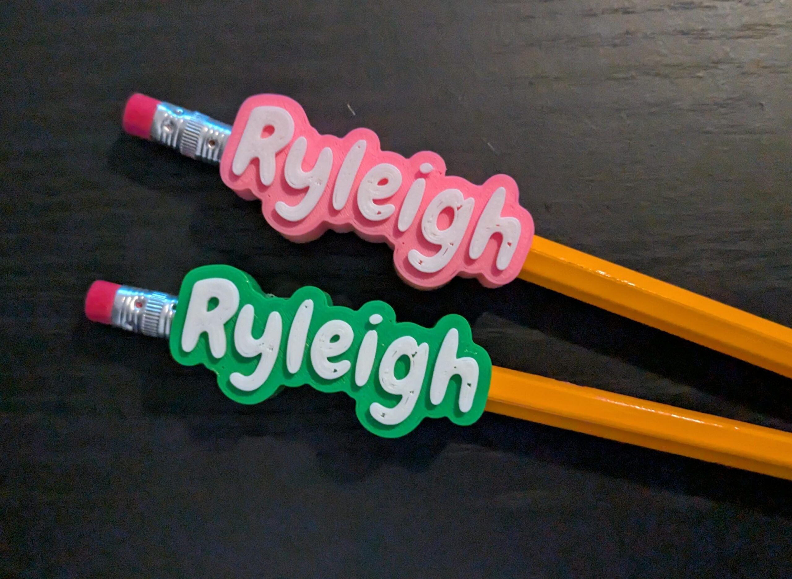 May include: Two yellow pencils with pink erasers and metal ferrules. Each pencil features a custom topper with the name Ryleigh in white letters. One topper is pink with a scalloped edge, and the other is green with a scalloped edge.