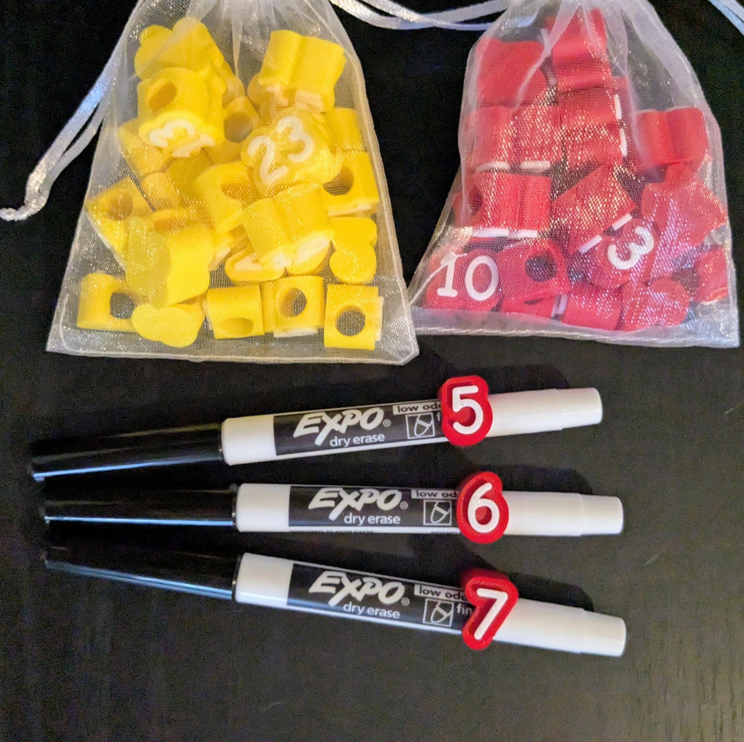 May include: Two sheer drawstring bags containing small yellow and red plastic number markers. Three white Expo dry erase markers with black caps are positioned below the bags, each featuring a red plastic number clip labeled 5, 6, and 7.