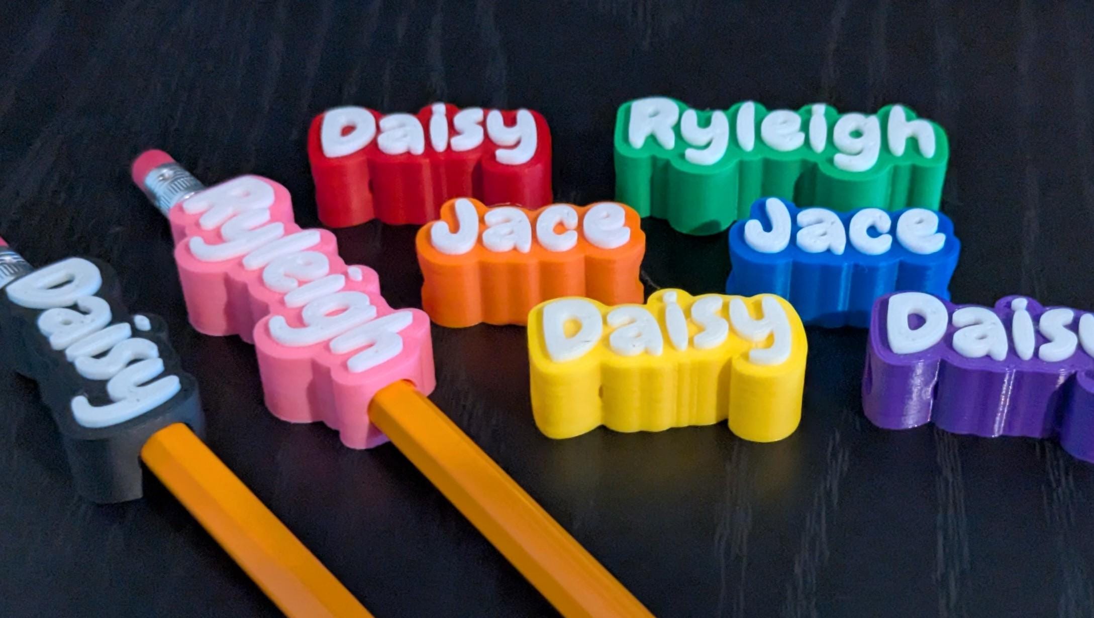 May include: A collection of colorful pencil toppers featuring the names Daisy, Ryleigh, and Jace in raised white lettering. The toppers are available in red, green, orange, blue, yellow, purple, and black, with two pencils inserted into the toppers.