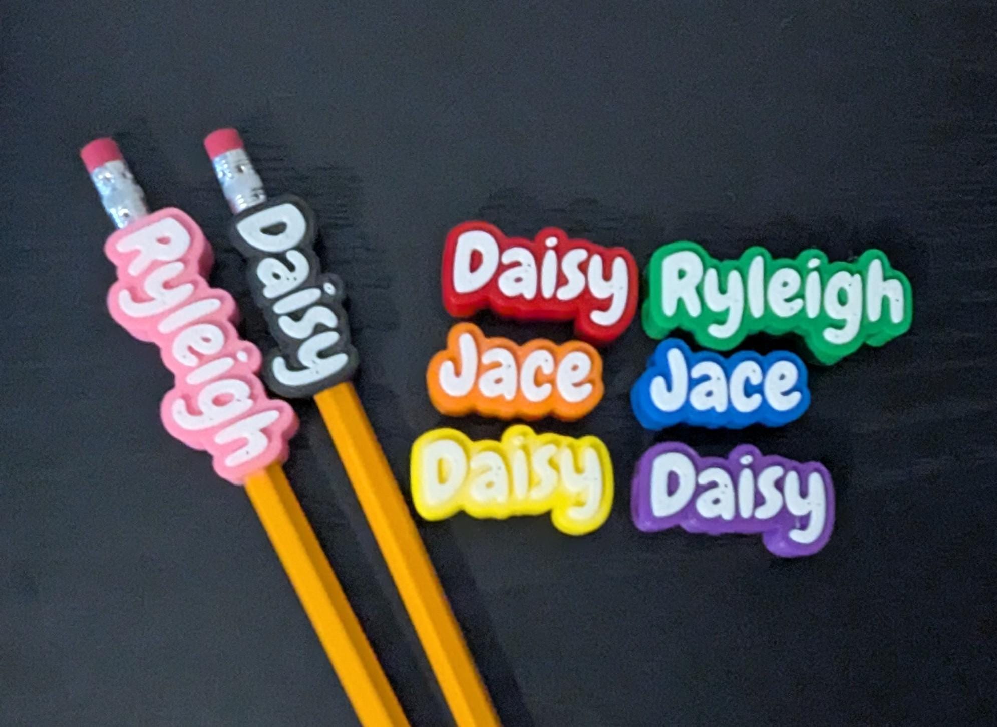 May include: Two yellow pencils with personalized silicone toppers featuring the names Ryleigh and Daisy in white text. Beside them are six additional silicone name tags in red, orange, yellow, green, blue, and purple, displaying the names Daisy, Ryleigh, and Jace.