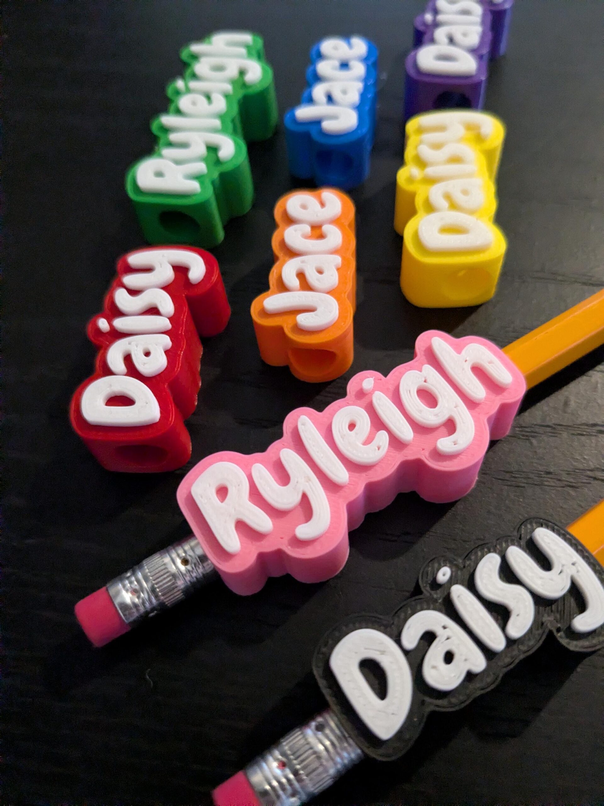 May include: A collection of colorful 3D-printed pencil toppers featuring raised white lettering. The names Daisy, Ryleigh, and Jace are displayed in various colors including green, blue, purple, yellow, orange, red, pink, and black on pencils.
