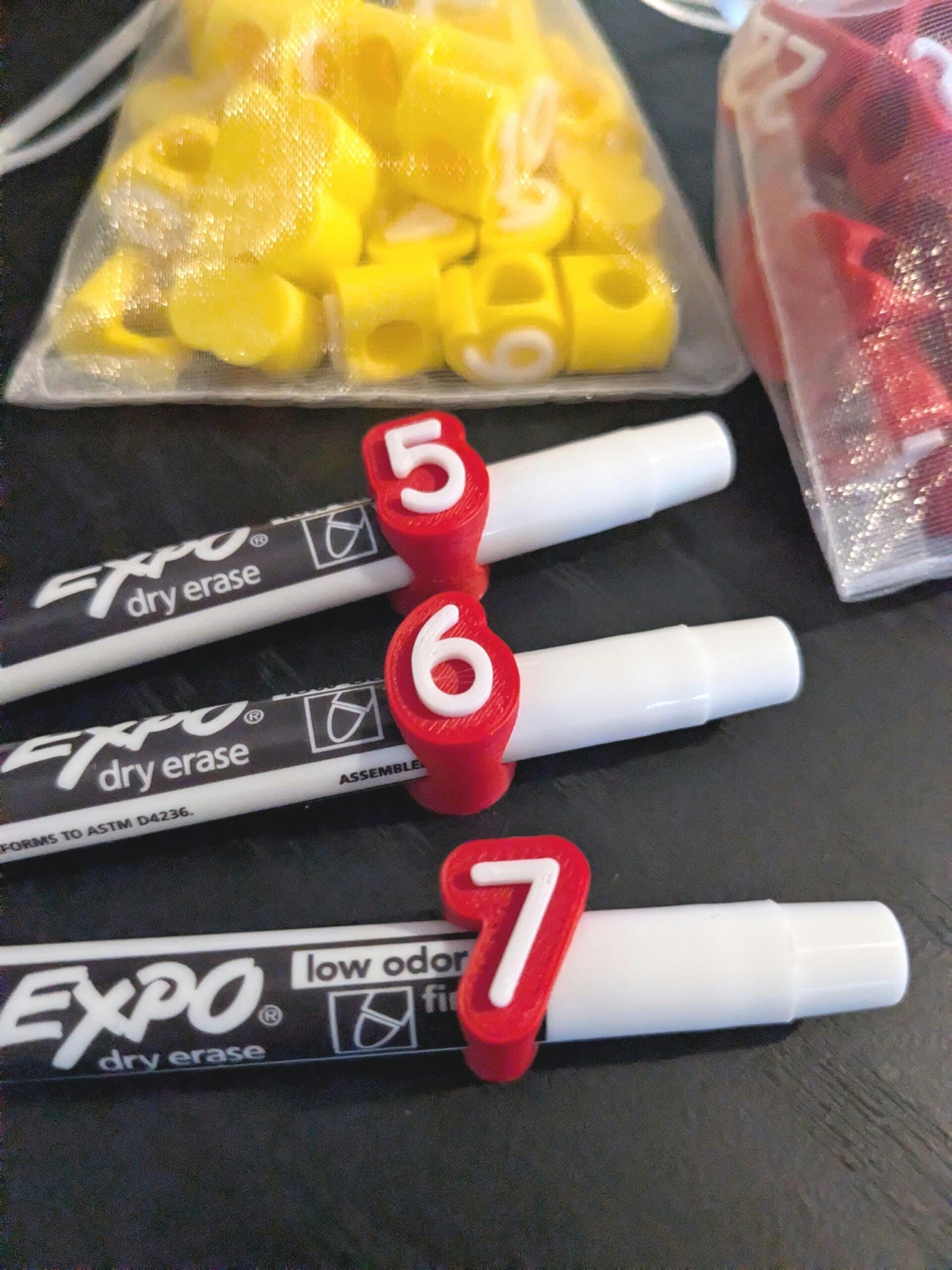 May include: Three black and white Expo dry erase markers, each fitted with a red plastic clip featuring a raised white number five, six, or seven. Behind the markers are two sheer mesh drawstring bags containing additional yellow and red plastic clips.