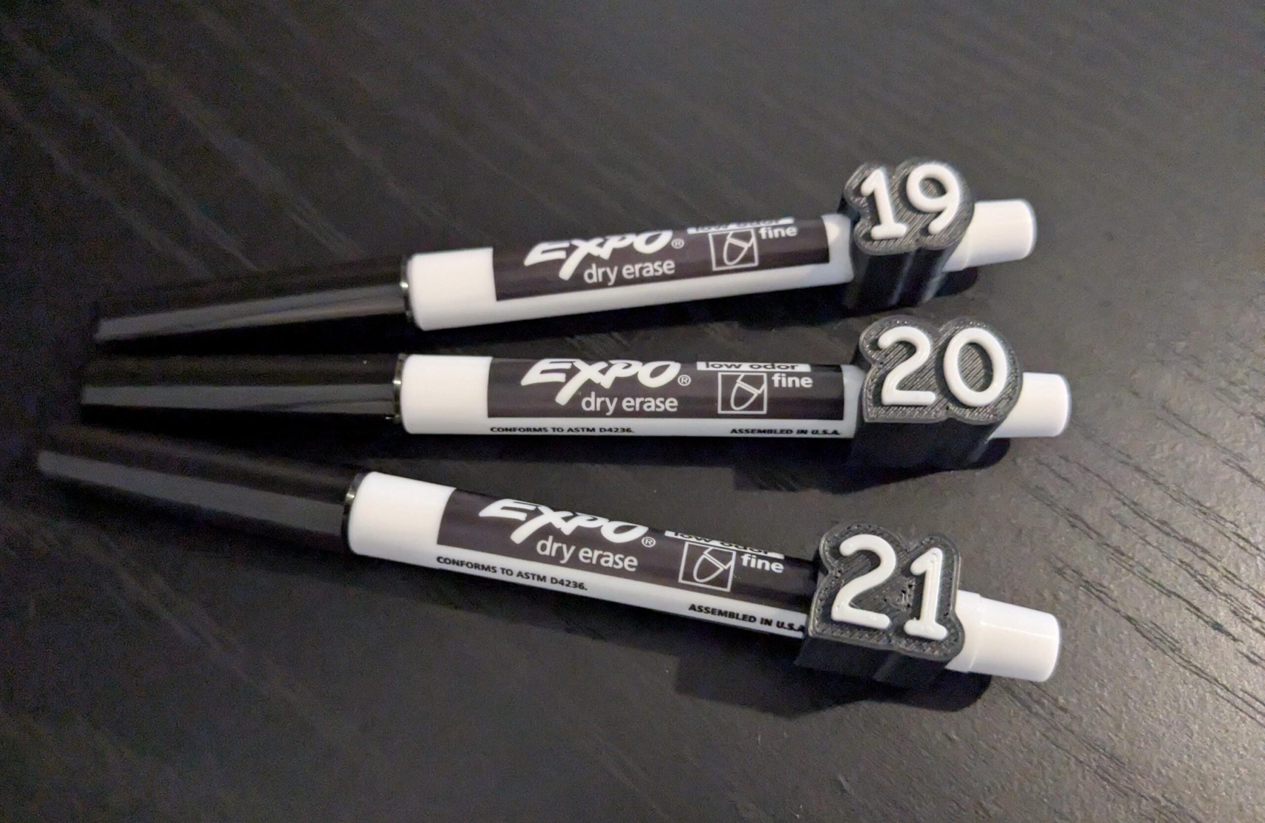 May include: Three black and white Expo fine point dry erase markers, each featuring a black 3D-printed plastic clip attached to the barrel. The clips are embossed with the white numbers 19, 20, and 21 respectively, identifying each individual marker.