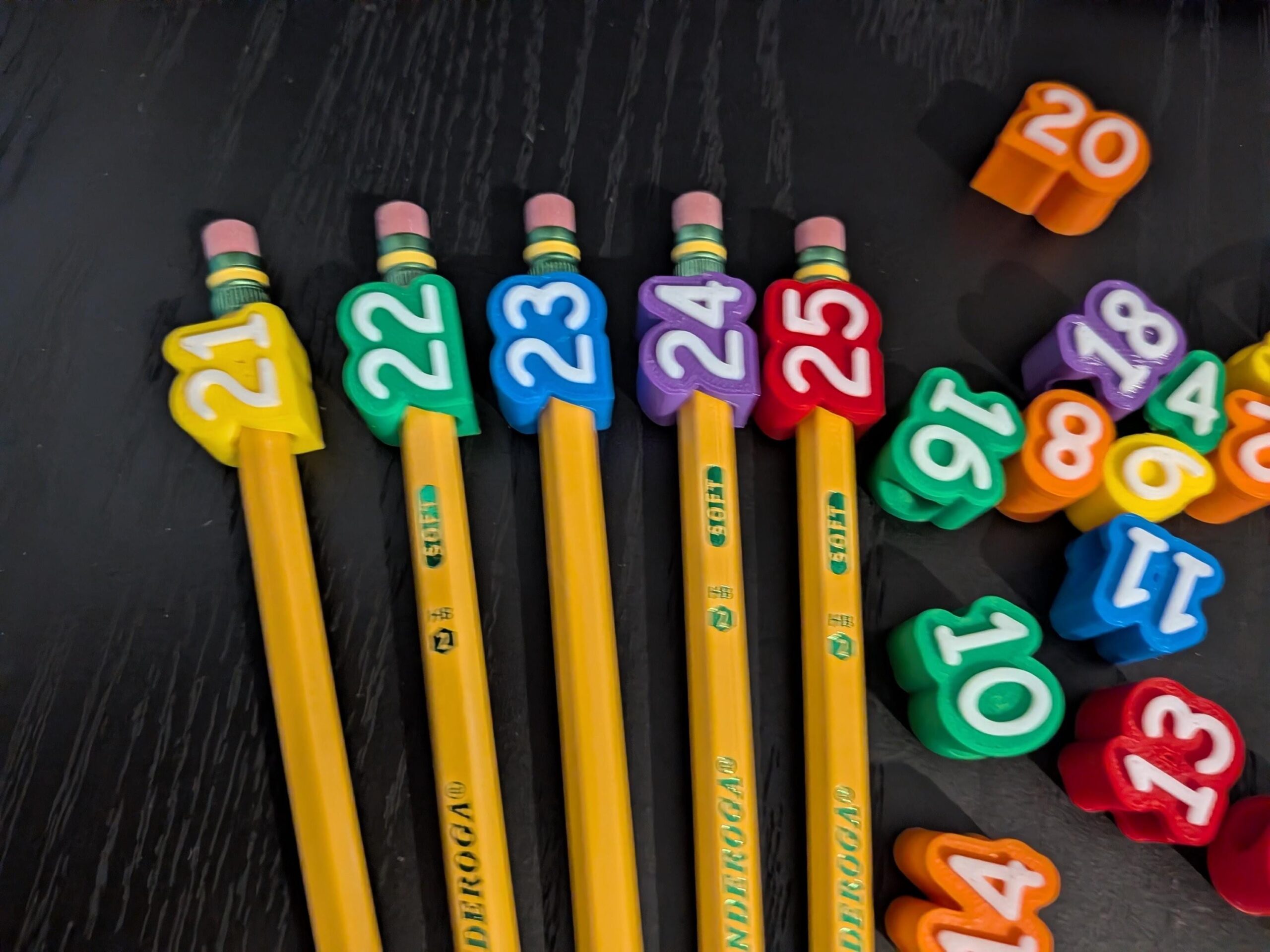 May include: Five yellow pencils with colorful number toppers 21, 22, 23, 24, and 25 attached to the erasers. A collection of loose, colorful number-shaped plastic toppers including 11, 13, 14, 16, 18, and 20 are scattered to the right side.