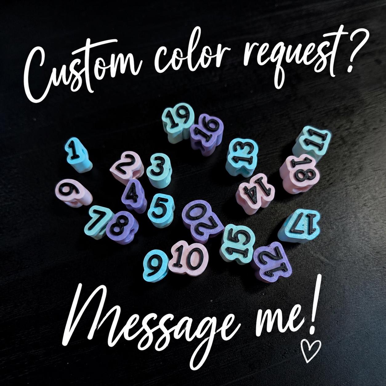 May include: A collection of small, shaped beads featuring the numbers 1 through 20 in black ink. The beads are colored in light blue and light purple. The text "Custom color request? Message me!" is displayed in white script above and below the beads.