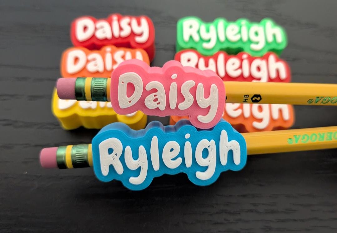 May include: Two yellow pencils with pink erasers and metal ferrules, each topped with a custom silicone name topper. One topper is pink with the white text Daisy, and the other is blue with the white text Ryleigh. Additional colorful name toppers are arranged behind.