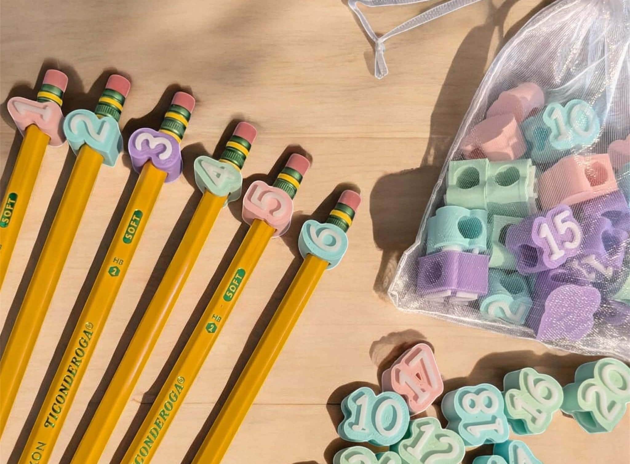 May include: Six yellow Ticonderoga pencils with pastel-colored number pencil toppers ranging from one to six, alongside a sheer white drawstring bag filled with additional pastel number toppers and loose toppers numbered ten through twenty.