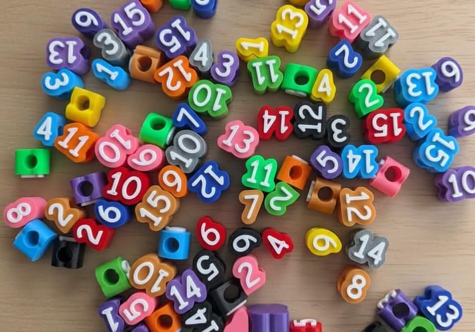 May include: A collection of small, multi-colored plastic beads, each shaped like a rounded square with a central hole. The beads feature white printed numbers ranging from 1 to 15, including colors like red, blue, green, yellow, orange, purple, and black.