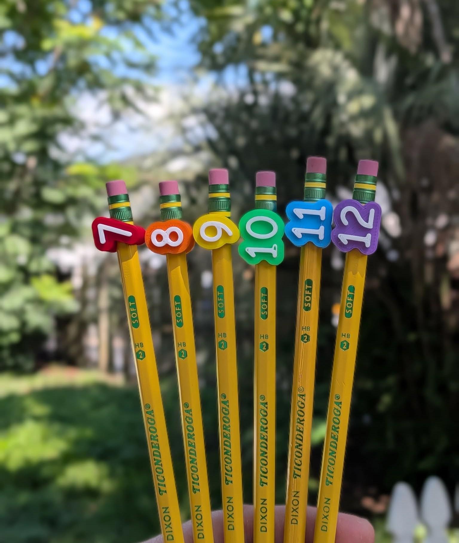 May include: Six yellow Dixon Ticonderoga pencils arranged in a row, each topped with a colorful rubber number topper ranging from seven to twelve. The pencils feature green metal ferrules with pink erasers and are marked with the text SOFT and HB 2.