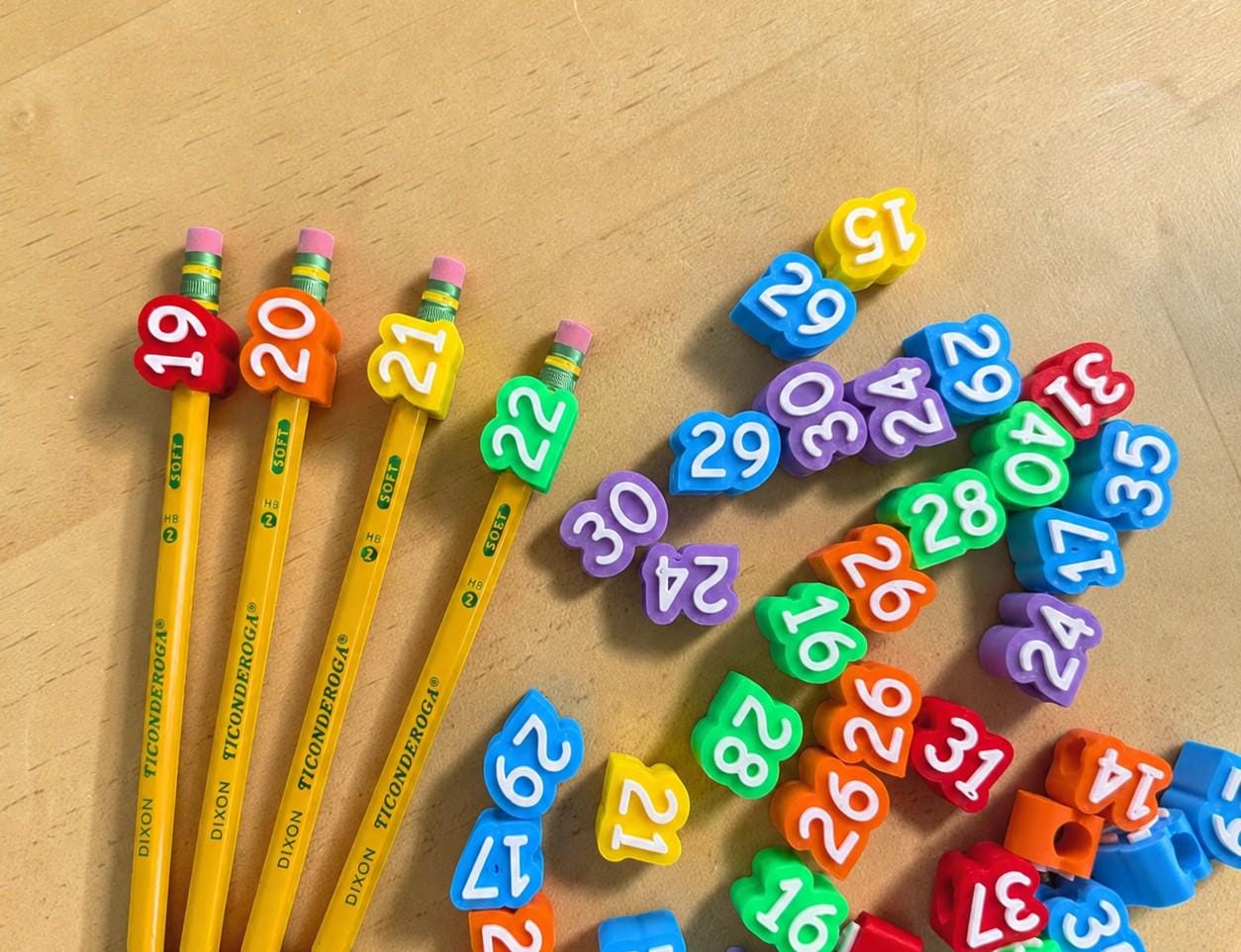 May include: Four yellow Dixon Ticonderoga pencils with colorful number-shaped rubber pencil toppers featuring the numbers 19, 20, 21, and 22. A collection of loose, multi-colored rubber number toppers is scattered nearby on the surface.
