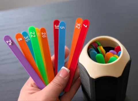May include: A set of colorful plastic sticks with white numbers printed on them, including 24, 8, 35, 22, 21, 26, 31, 5, 32, and 7. The sticks are held in a fan shape, and a matching black and beige pencil-shaped container holds more sticks.
