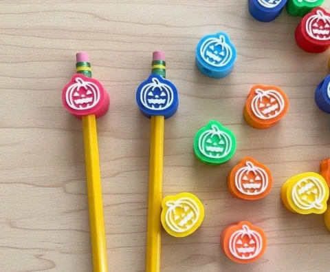 May include: A collection of pencil toppers shaped like round pumpkins in various colors including pink, blue, green, orange, and yellow. Each topper features a white jack-o'-lantern face design. Two yellow pencils are inserted into a pink and a blue topper.