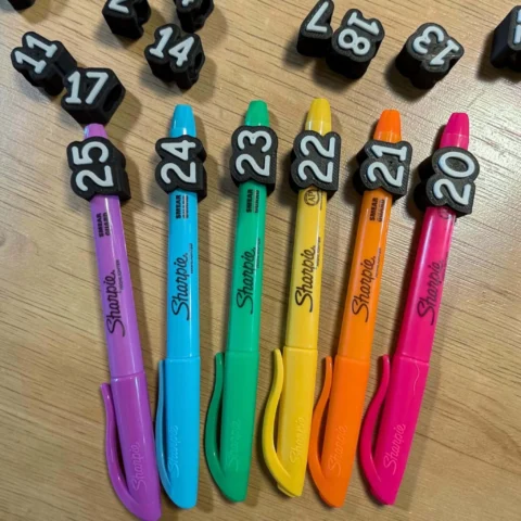 May include: Six Sharpie highlighters in purple, blue, green, yellow, orange, and pink, each featuring a black clip with a white number. The numbers 20, 21, 22, 23, 24, and 25 are attached to the pens, with loose black number beads nearby.
