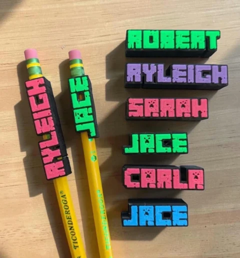 May include: Two yellow Ticonderoga pencils with pink erasers and custom 3D-printed name toppers in pink and green. Beside them are six rectangular name tags in black with block-style lettering in green, purple, pink, and blue colors.